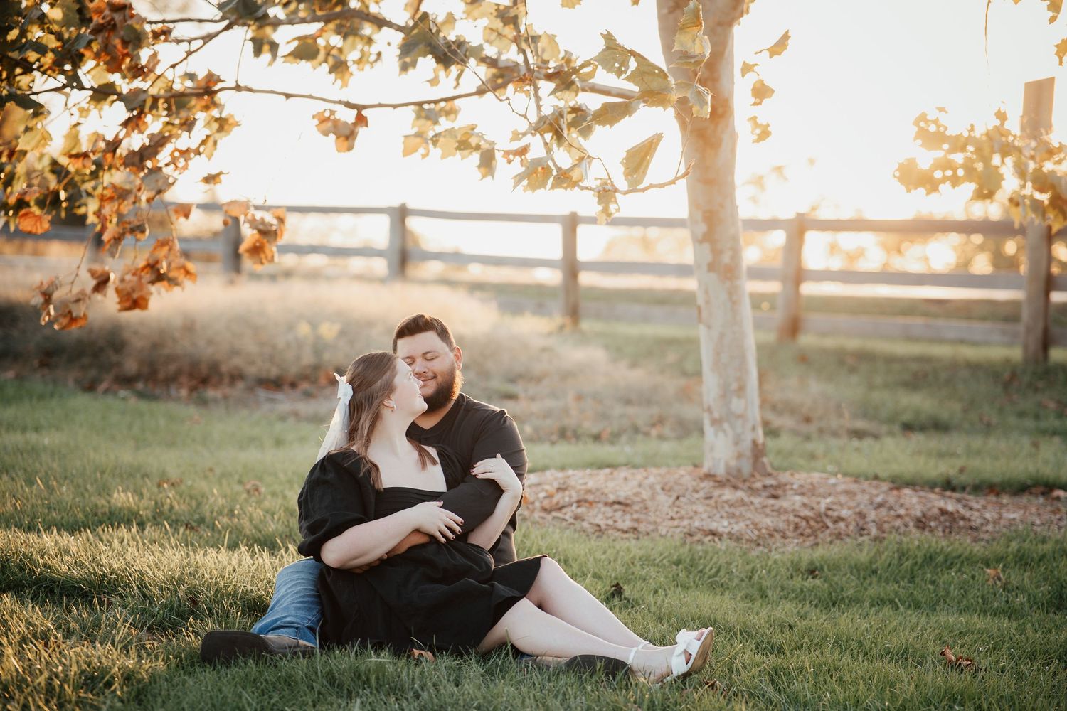 Couple sitting together on grass at sunset with wooden fence and autumn leaves in background during golden hour.