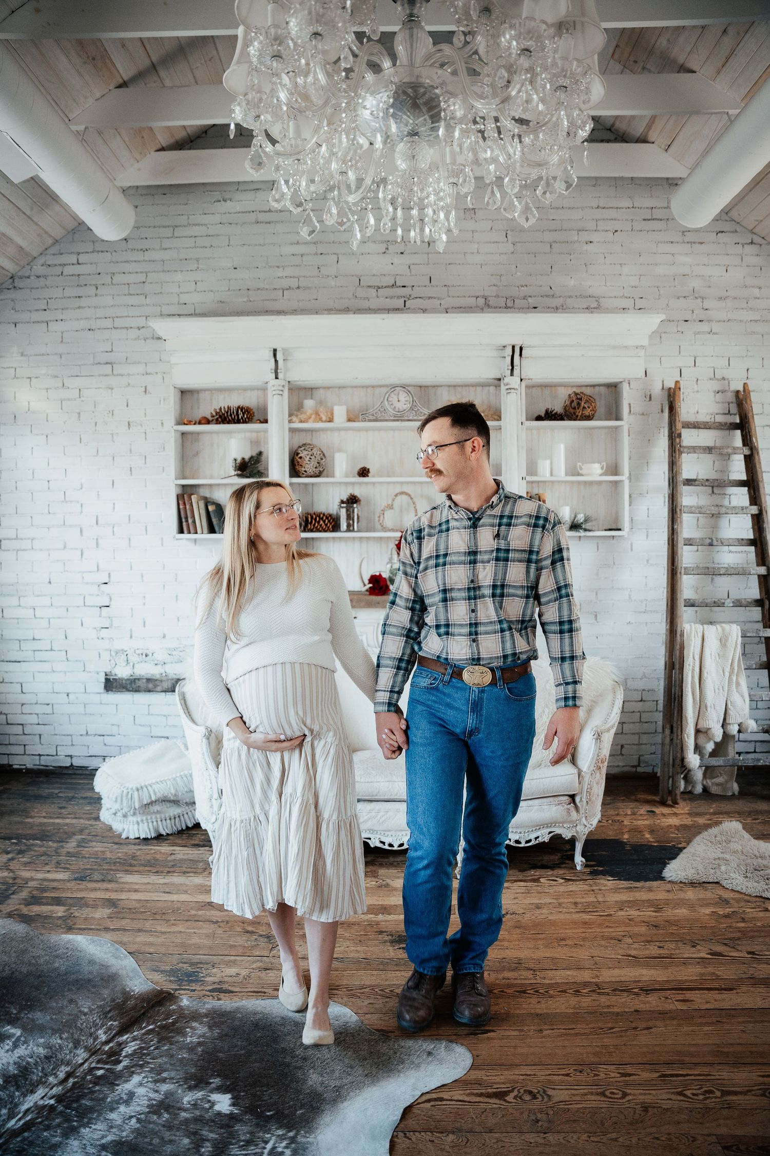 A couple poses in a bright white room with chandeliers and rustic wooden floors.