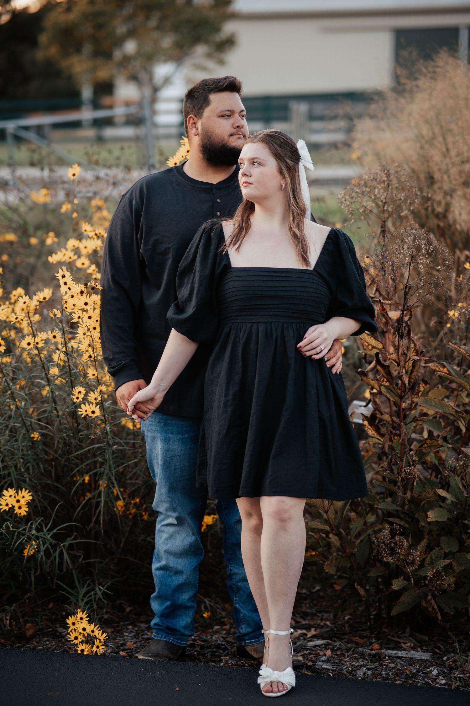 A romantic couple in coordinating black outfits pose together among yellow wildflowers at sunset.