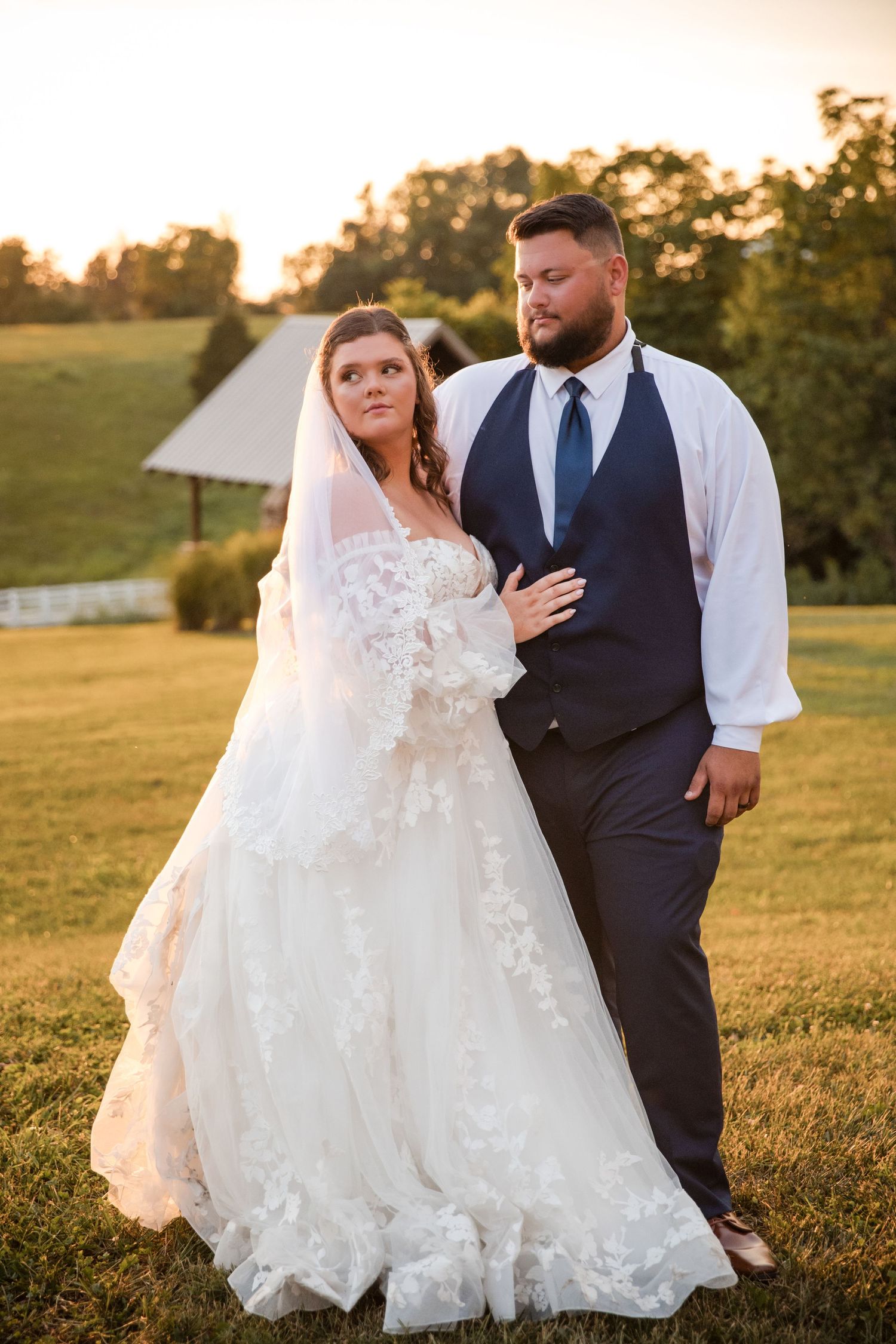 Bride in lace gown and veil stands with groom in navy vest and blue tie at golden hour outdoor wedding.