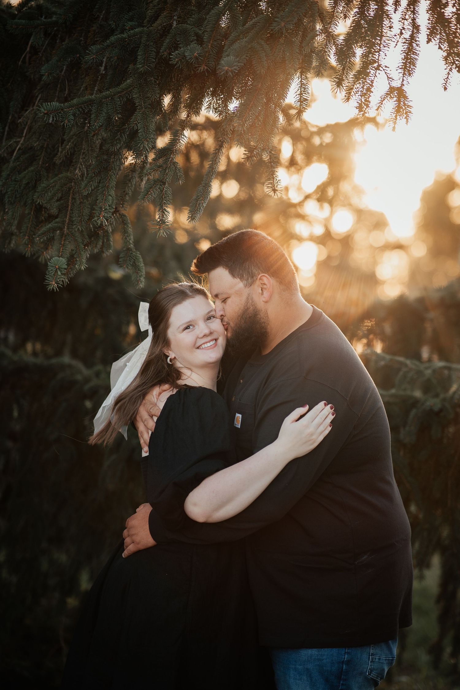 A romantic couple embraces during golden hour sunset under evergreen trees creating a dreamy backlit silhouette.