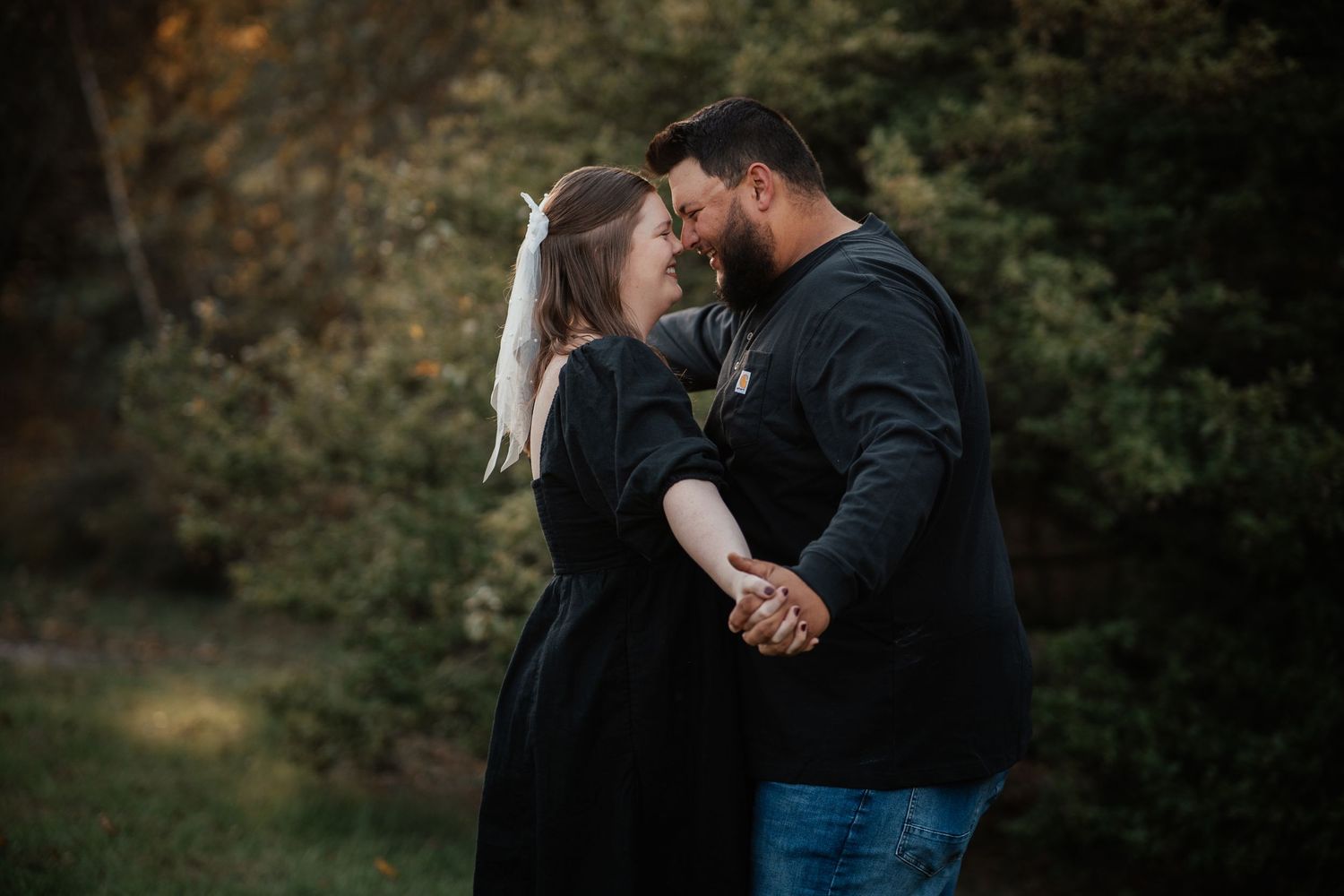 Two people in black clothing stand close together in an intimate embrace against a blurred natural outdoor background.