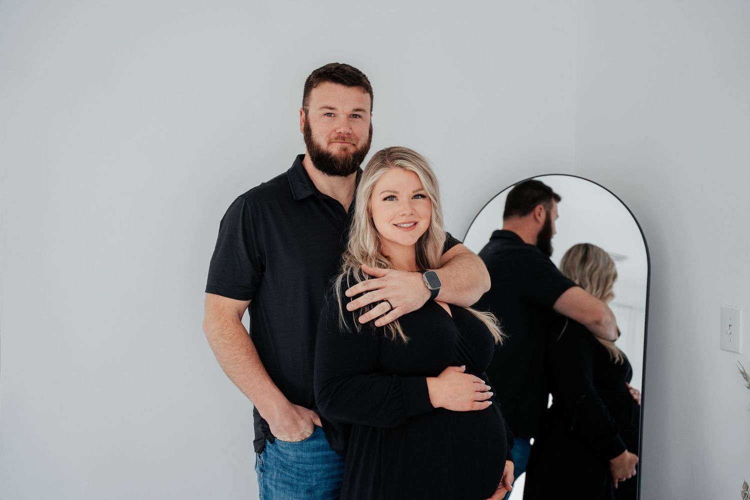 A couple in black clothing poses together against a white wall with a mirror reflection.