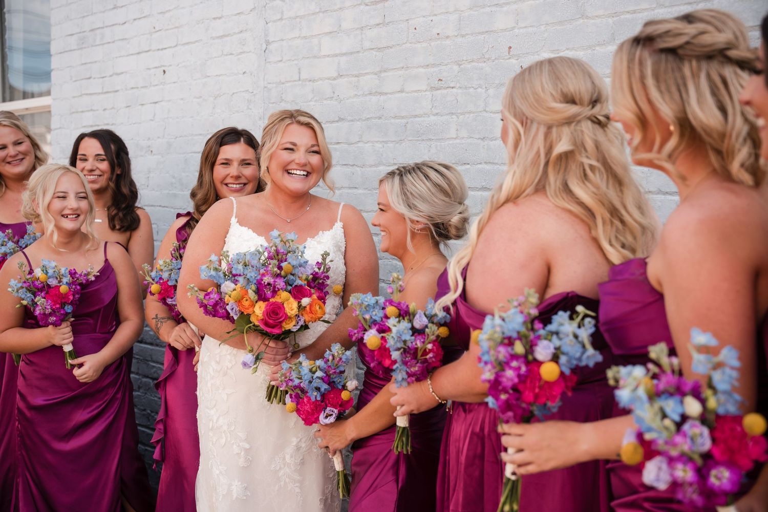 Bride in white gown smiles with bridesmaids in purple dresses holding colorful bouquets at outdoor wedding.