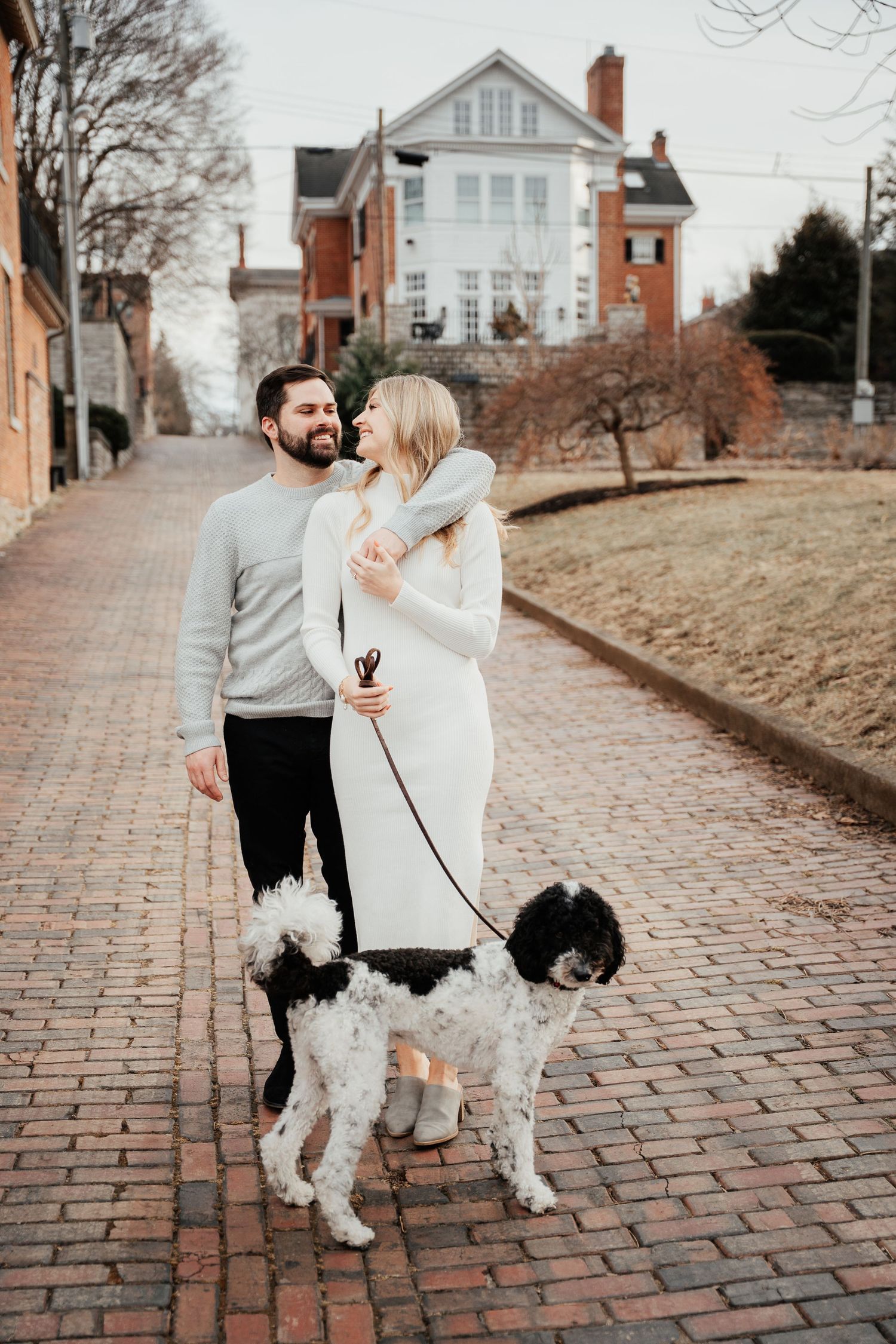 A couple in matching cream sweaters walks their black and white dogs on a brick path in front of a historic mansion.
