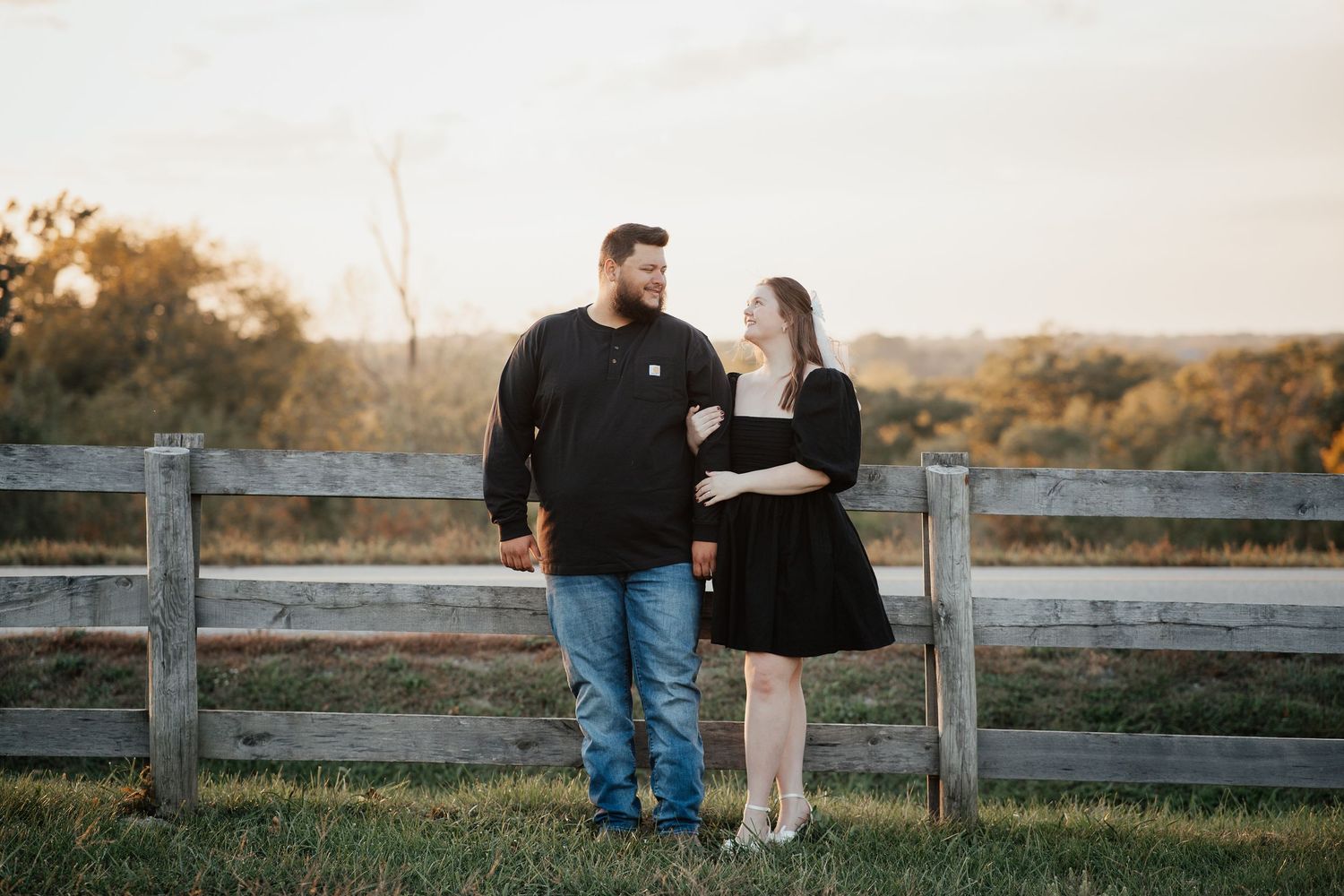 A couple shares a tender moment together while leaning against a wooden fence at sunset in a rural setting.