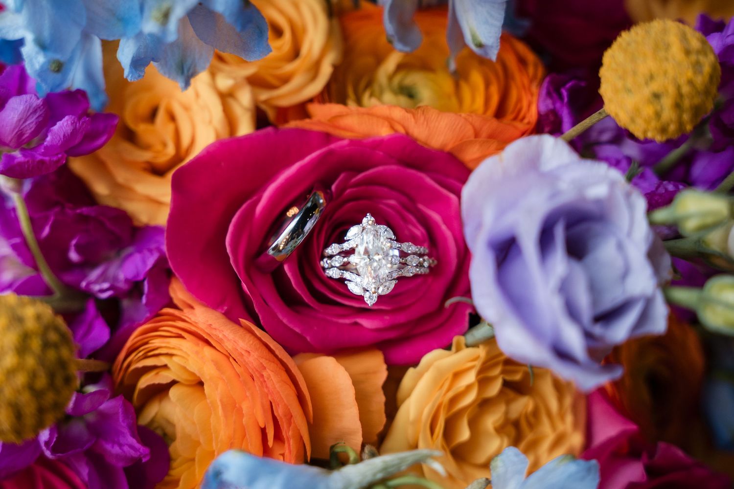 Diamond engagement ring and wedding band nestled in vibrant pink rose surrounded by colorful orange, purple, and blue flowers.