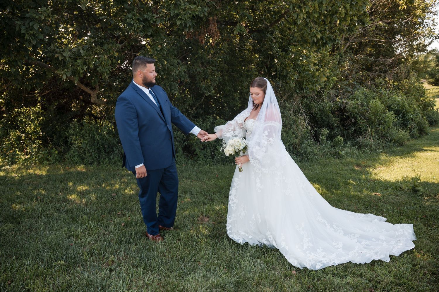A bride in a lace gown and veil holds hands with a groom in a navy suit outdoors on green grass.