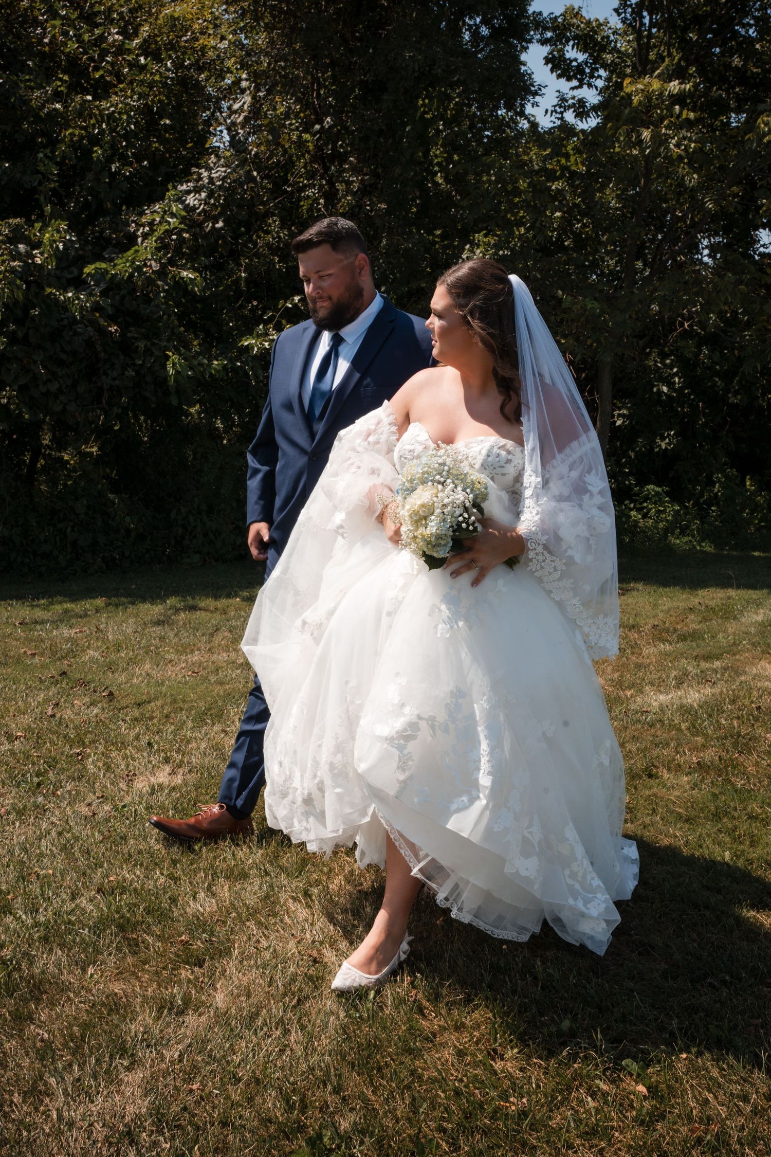 A bride in a white lace gown holds a bouquet while walking with a groom in a navy suit on a sunny lawn.