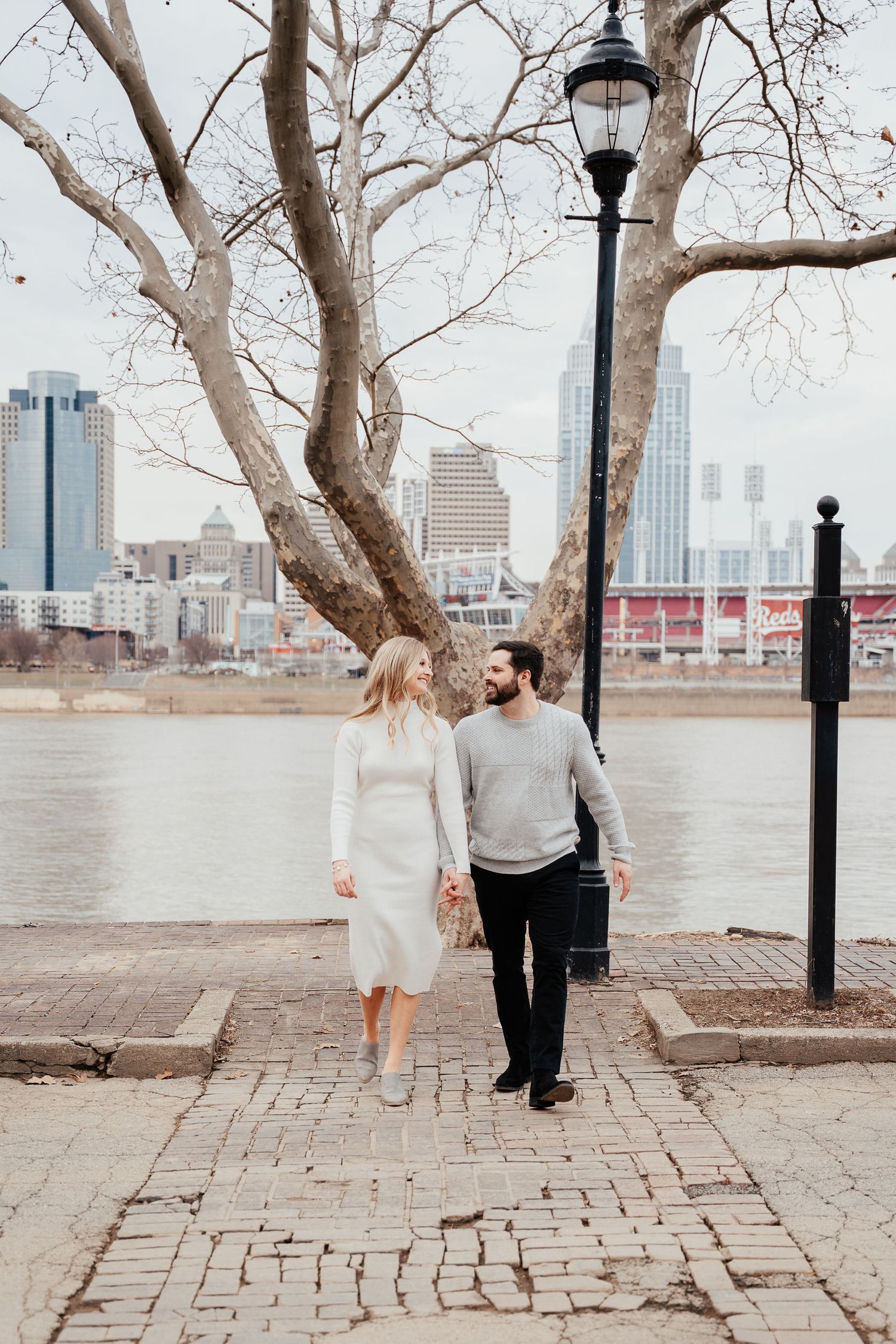 A couple strolls along a brick pathway near a lamppost with the Cincinnati skyline visible in the background.