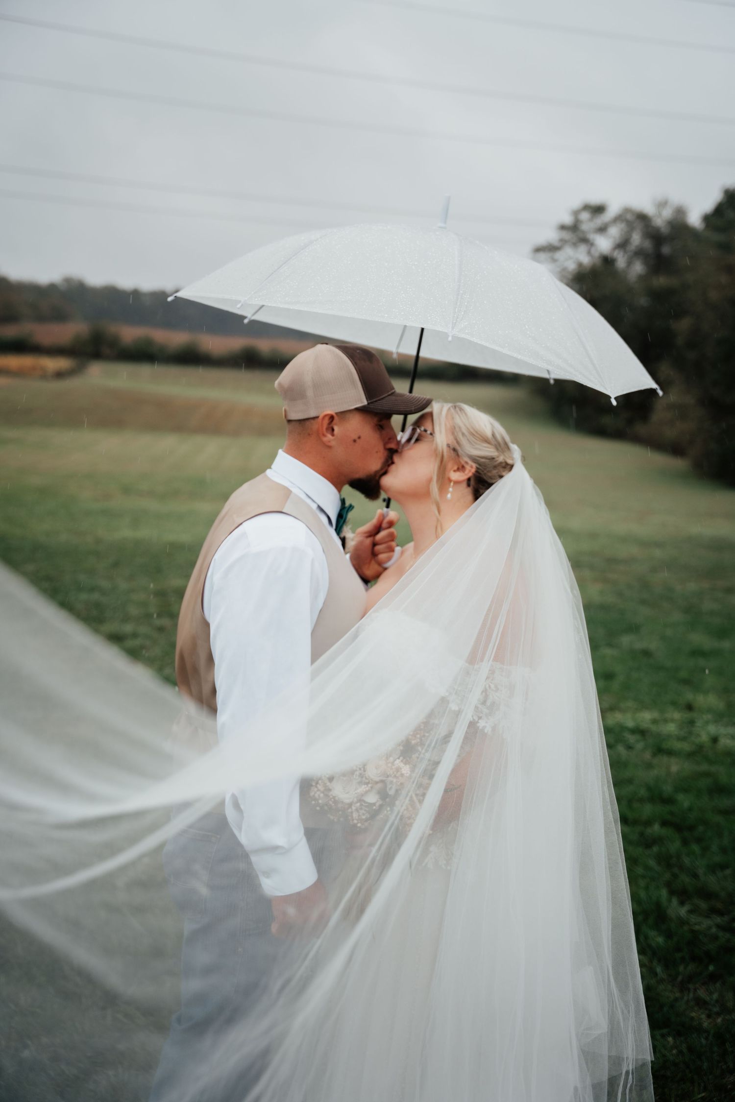 A romantic wedding moment shared under a white umbrella with a flowing veil in a countryside setting.