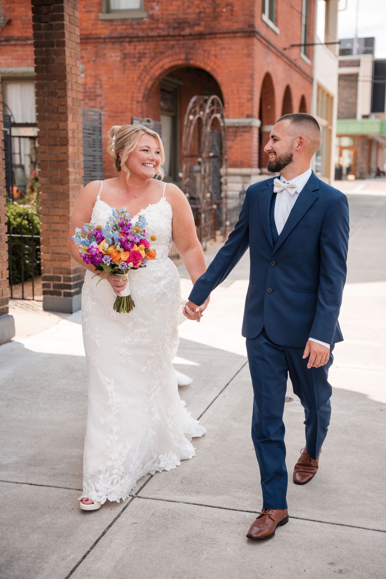 A bride and groom walk hand-in-hand on a city sidewalk, she in lace gown holding colorful bouquet, he in navy suit.