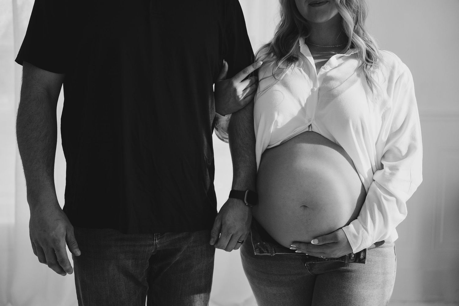 Black and white maternity photo showing couple holding hands with woman wearing white button up shirt revealing pregnant belly.