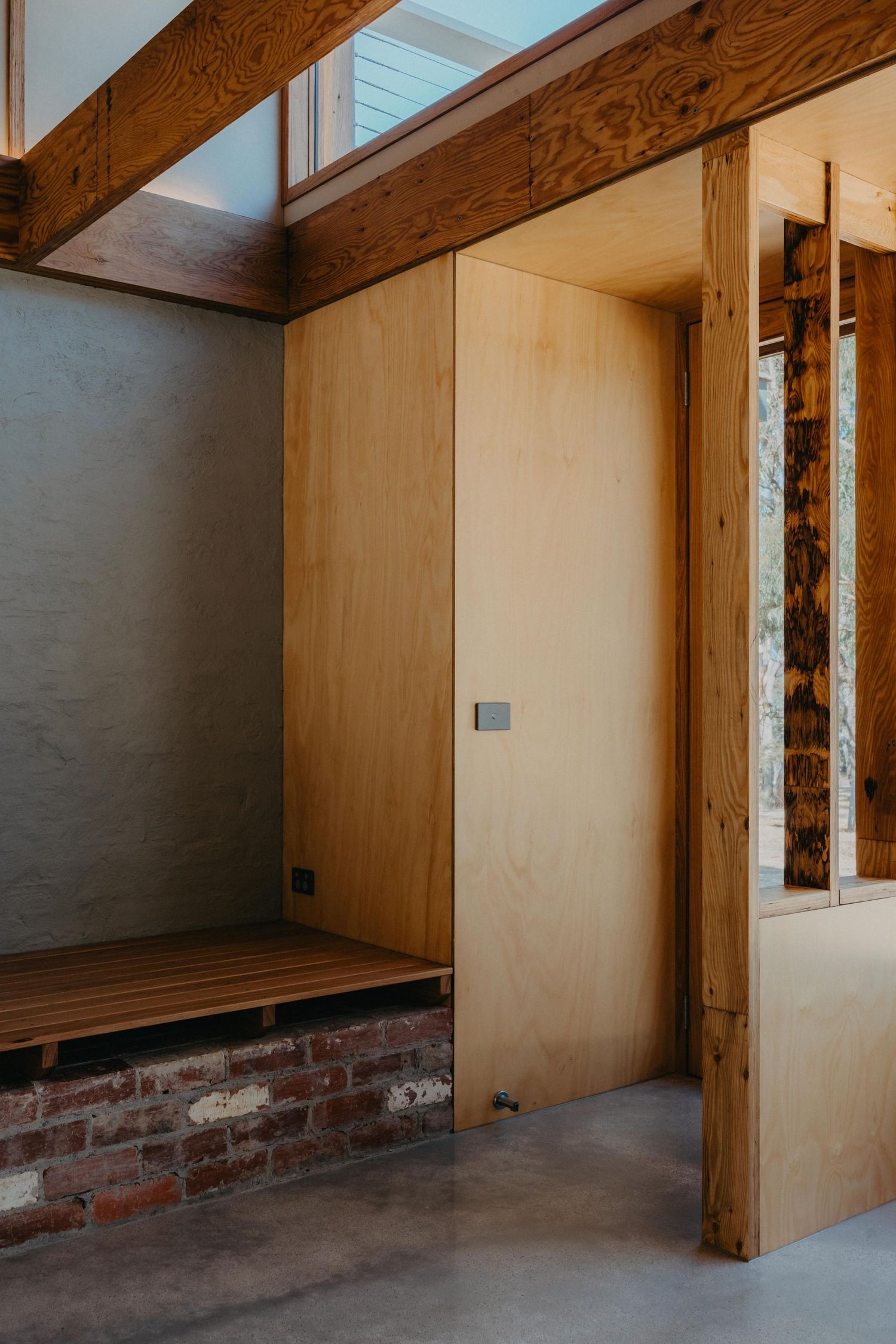 Plywood interior wall with timber bench on brick base, exposed beams, and concrete floor.