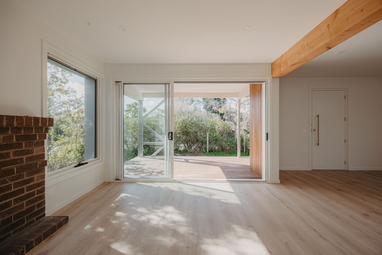 Sunlit open plan living space with exposed beam, brick fireplace, and garden view glass doors.