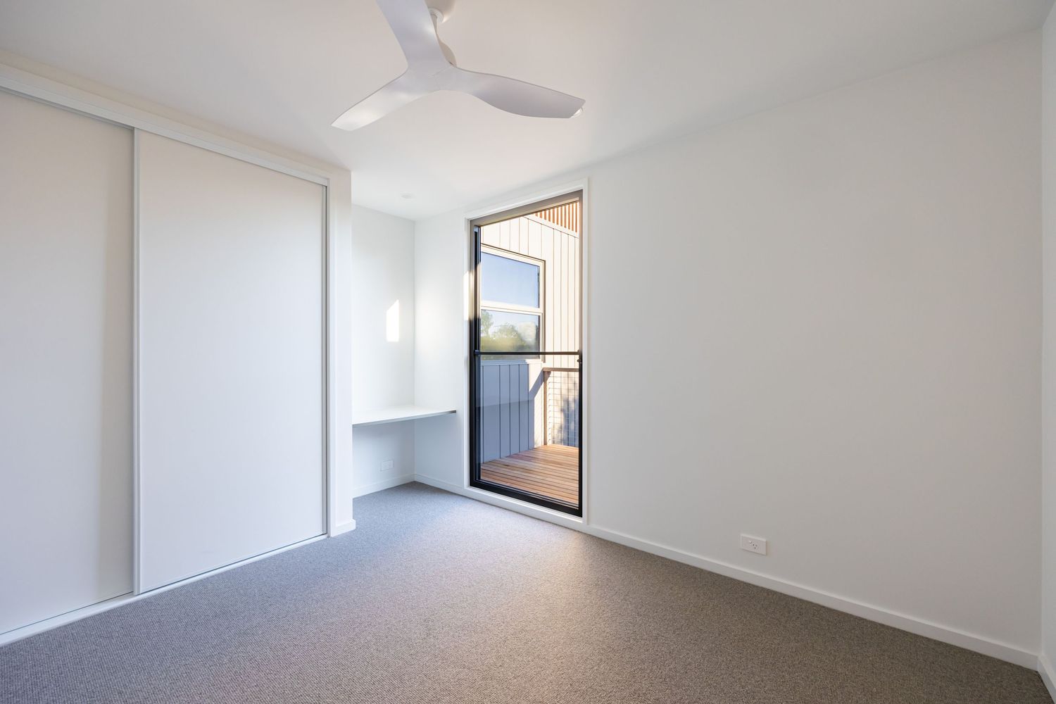 Bright empty bedroom with sliding wardrobe, ceiling fan, and glass door opening to a small balcony with timber decking.
