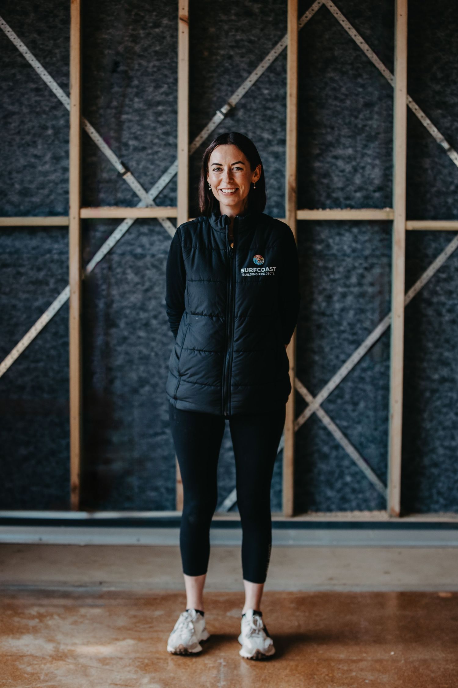Smiling woman in black vest and leggings standing confidently in a construction site interior.