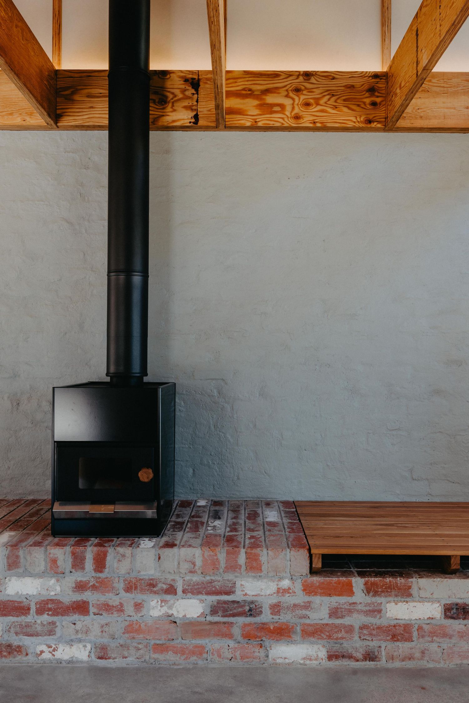 Black wood-burning stove on red brick hearth with timber bench beside grey rendered wall and exposed wooden ceiling beams.