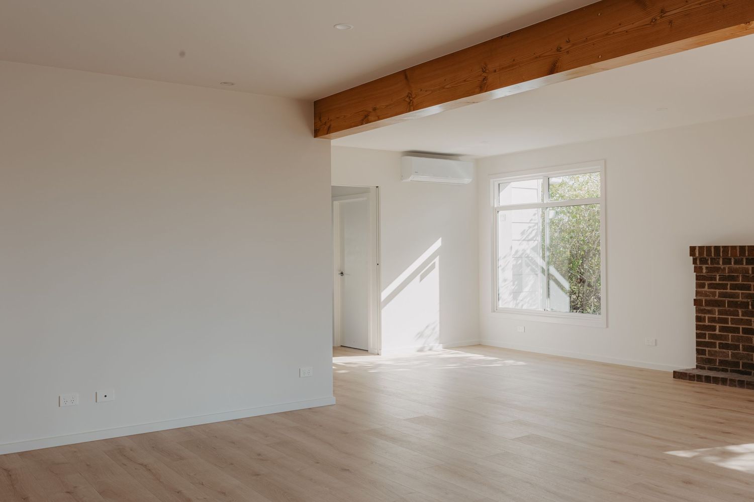 Bright minimalist living room with exposed timber beam, light hardwood floors, and brick fireplace.