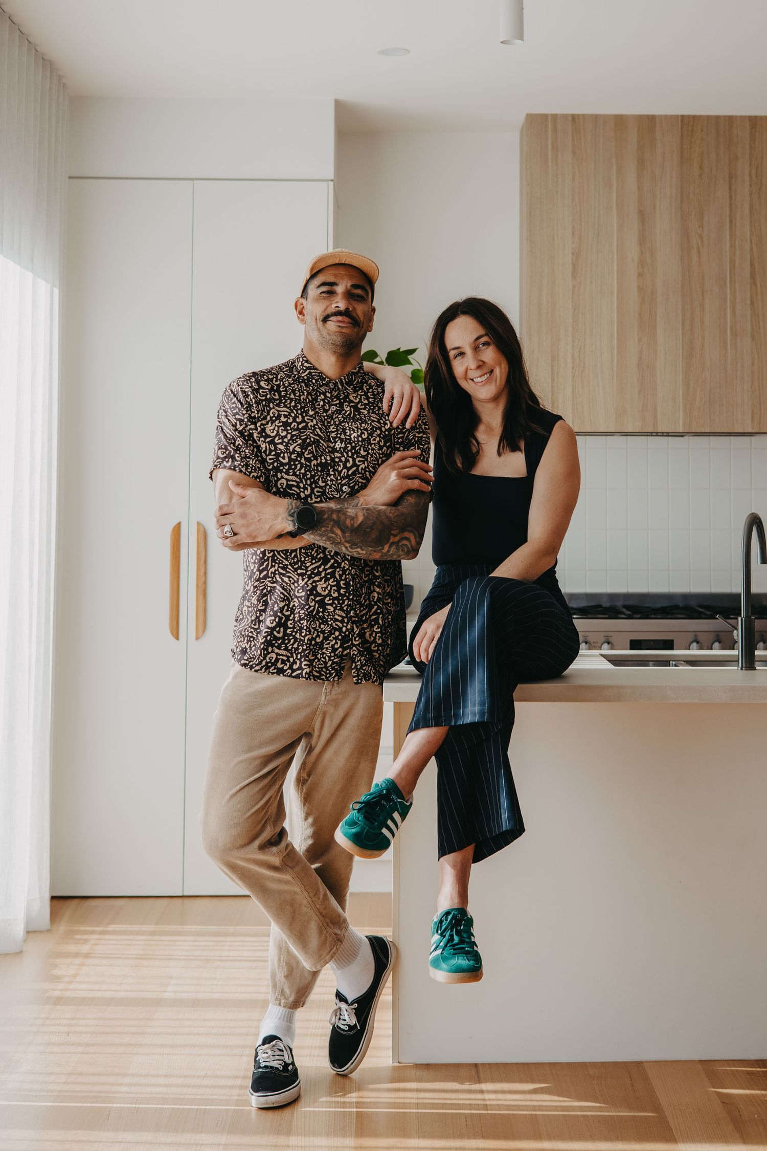 A stylish couple poses in a modern kitchen with timber accents, wearing casual streetwear and colorful sneakers.