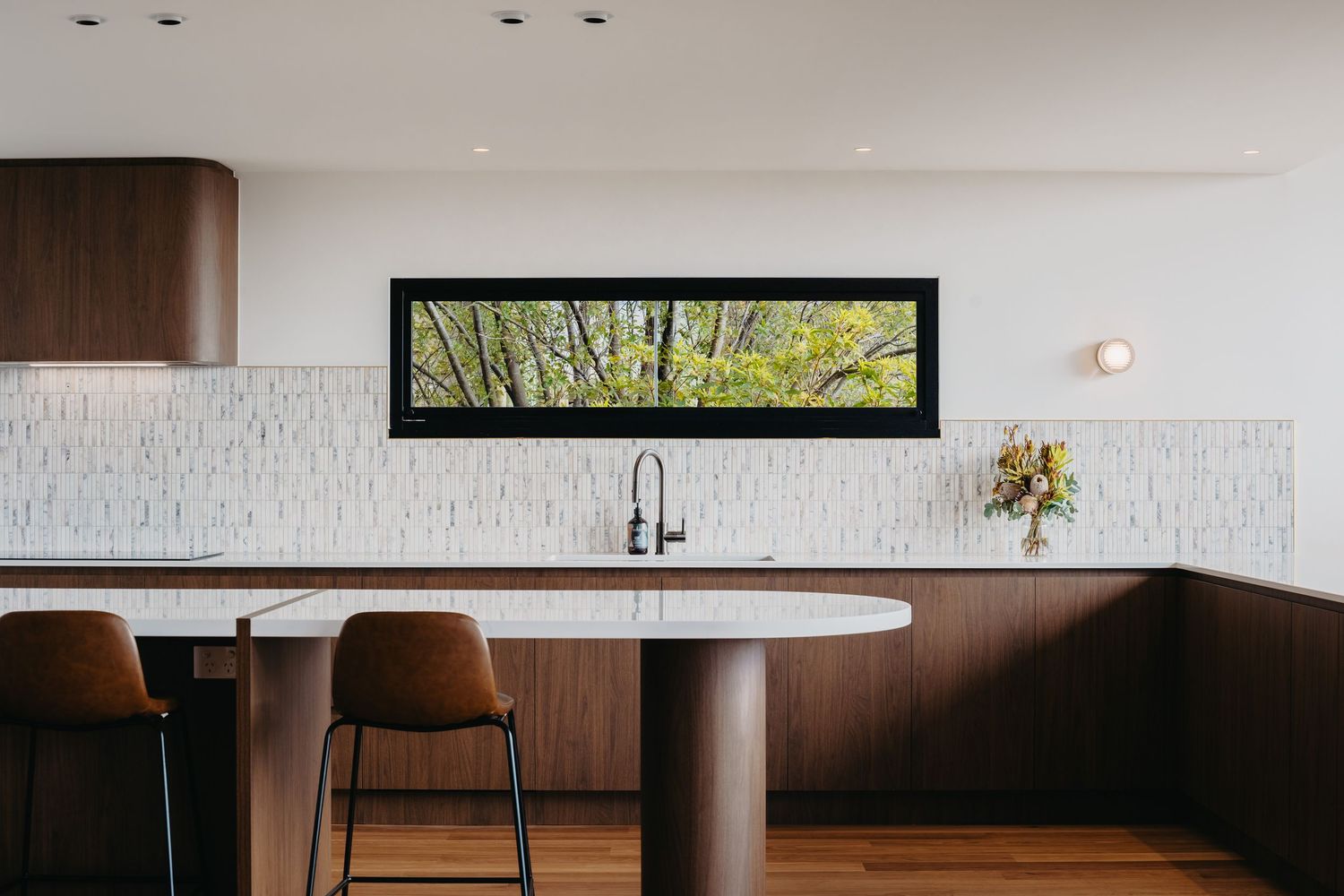 Modern kitchen with walnut cabinets, white stone island, bar stools, textured tile backsplash and panoramic window framing green trees.