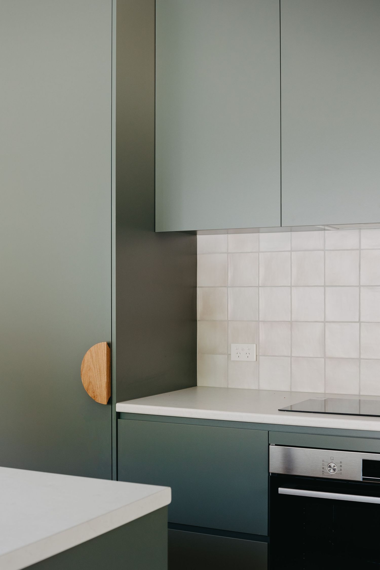 Modern kitchen with sage green cabinets, white square tile backsplash, round wooden handle, and built-in black oven.
