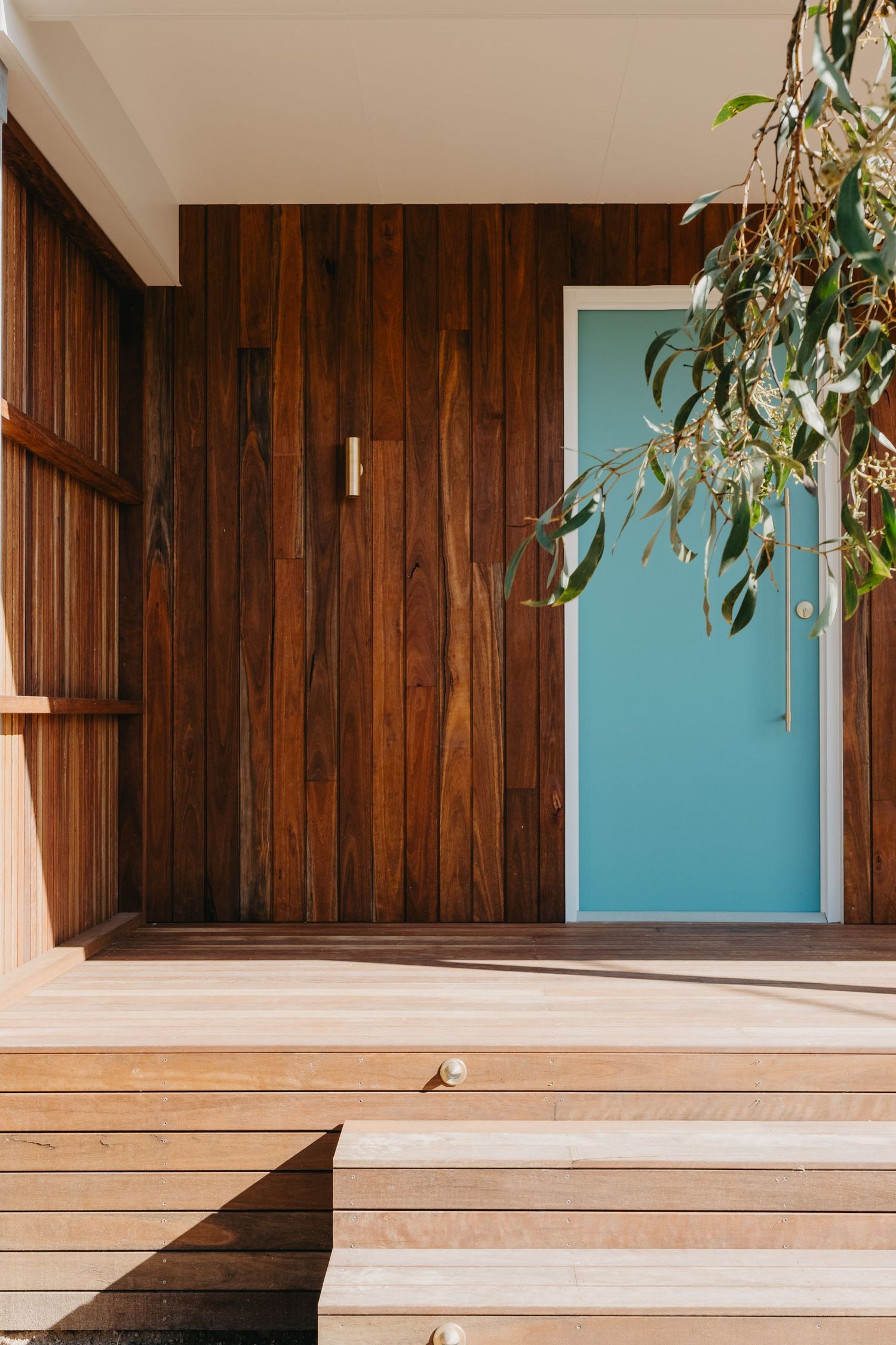 Timber-clad entrance with warm wood paneling, light wood deck steps, and a striking teal door surrounded by lush greenery.