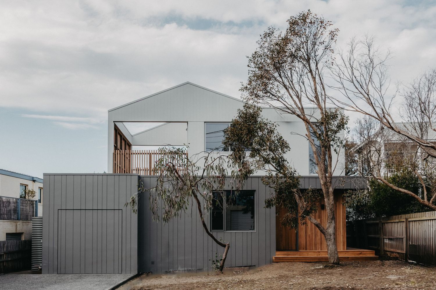 Modern grey corrugated steel home with timber deck, large trees, and industrial-style gate under overcast sky.