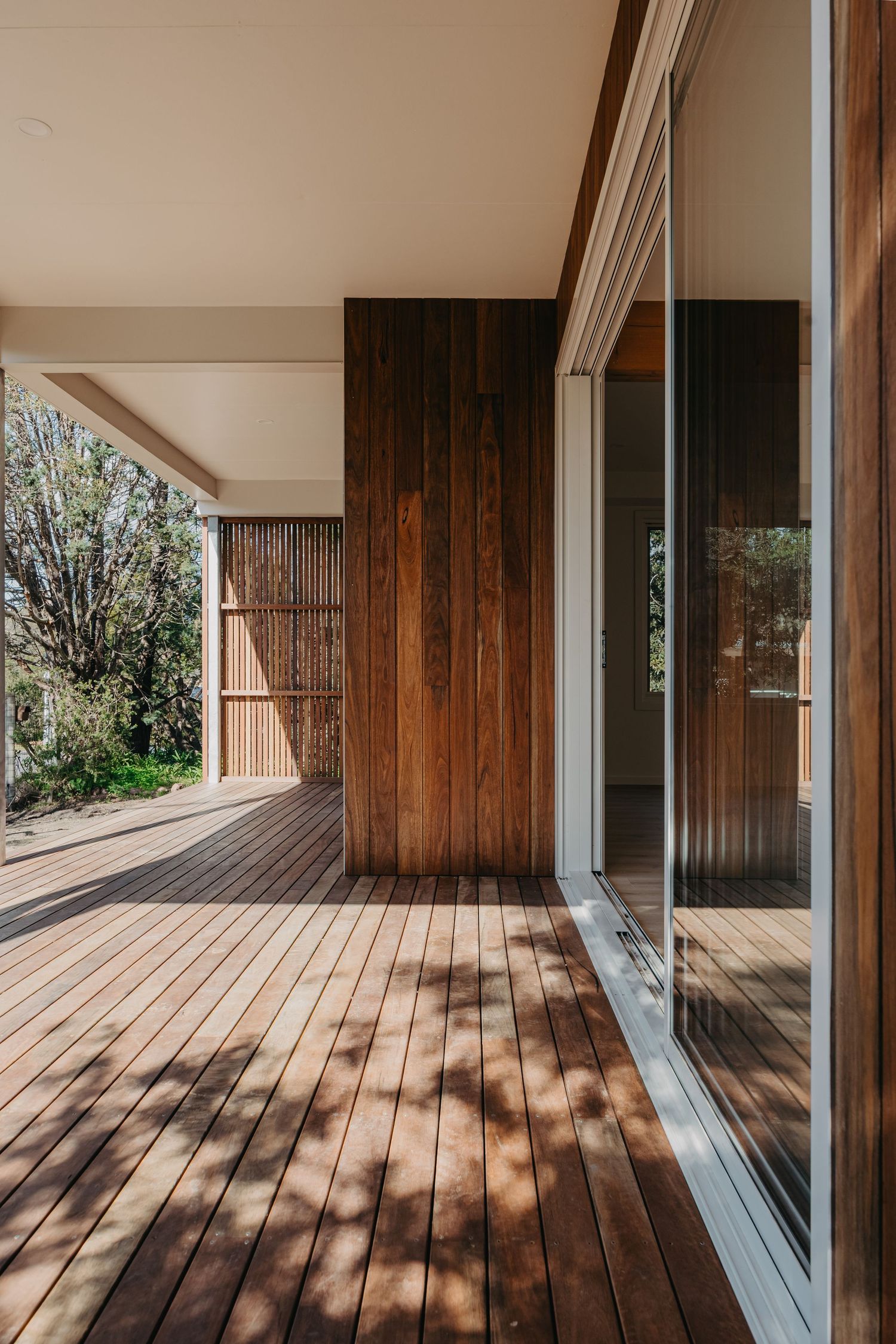 Sunlit timber deck with warm wood vertical cladding, large glass sliding doors, and dappled tree shadows.