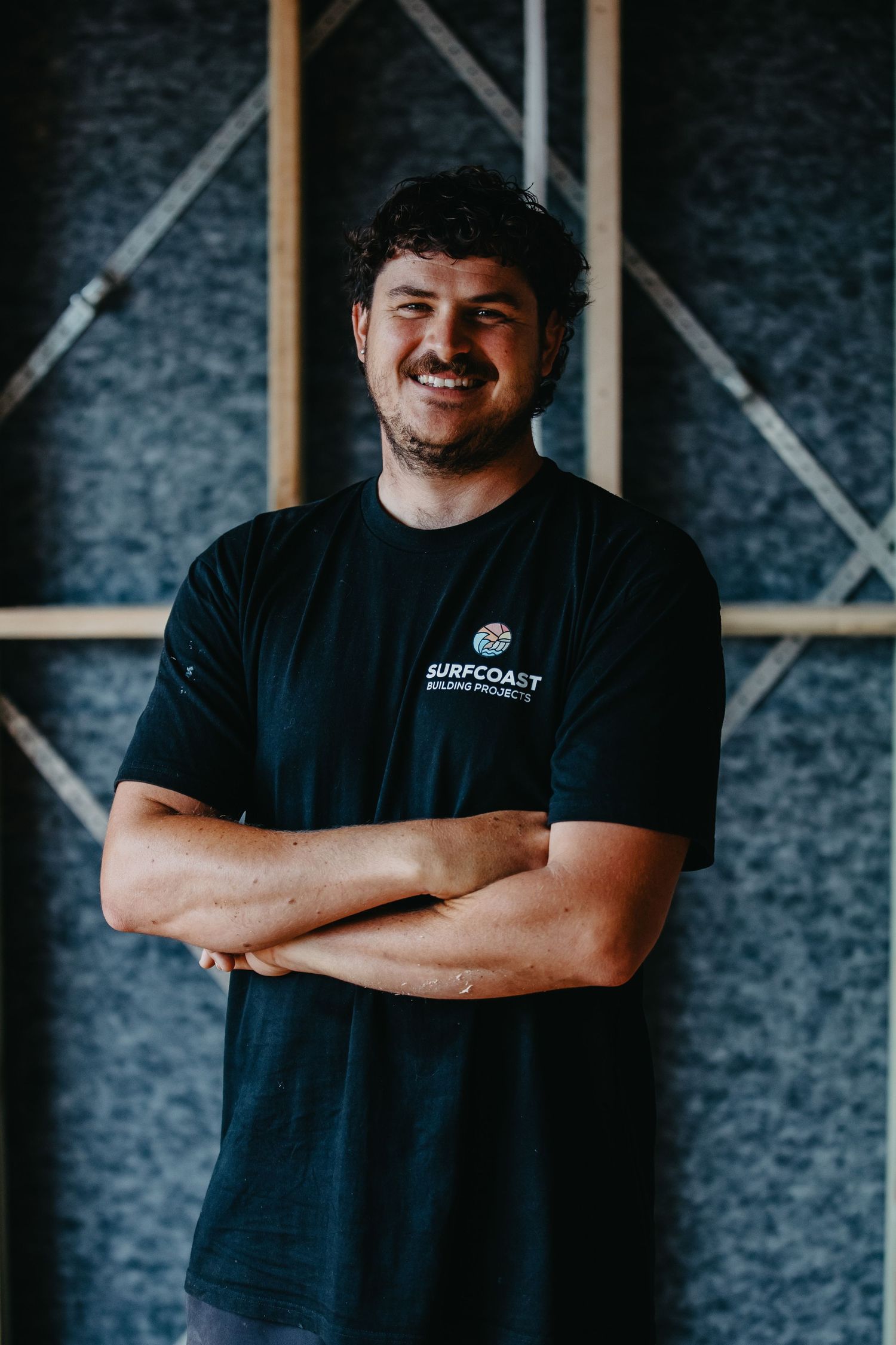 Man in black Surfcoast Building Projects t-shirt smiling with arms crossed in a modern interior space.