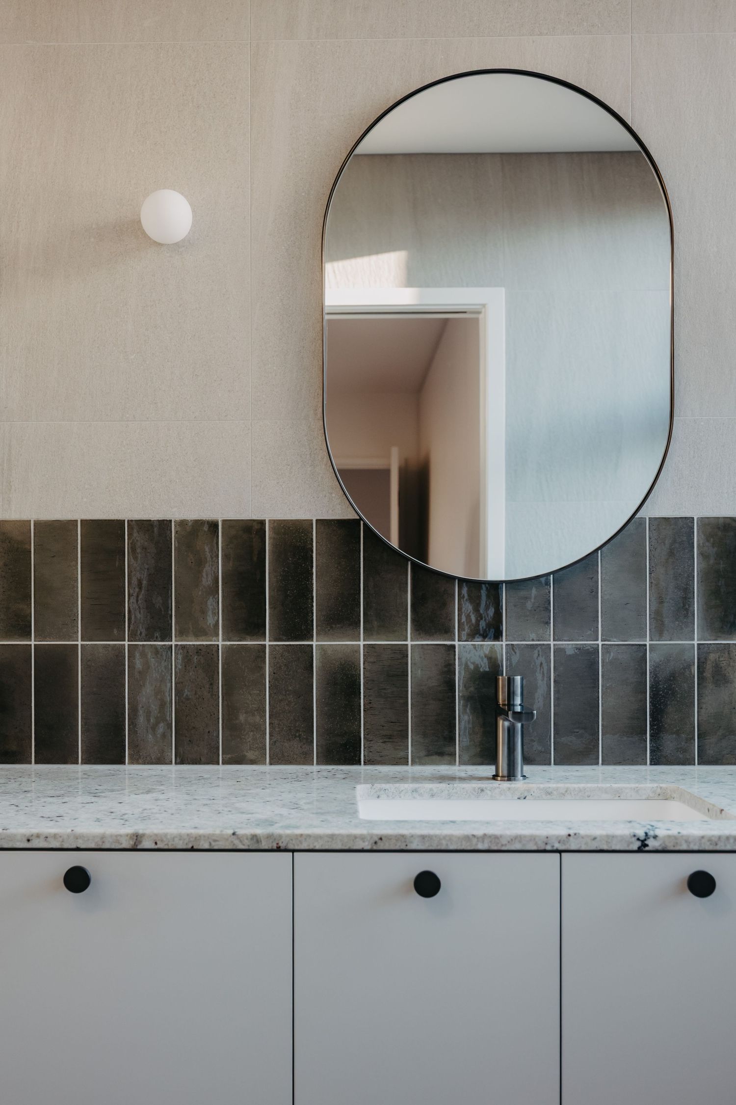 Modern bathroom vanity with oval black-framed mirror, dark vertical tiles, marble benchtop, and white cabinetry with round handles.
