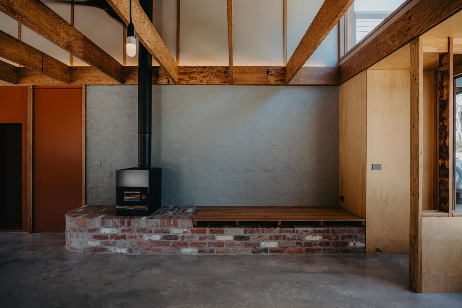 Modern interior with black freestanding wood heater on brick plinth, exposed timber rafters, plywood walls and polished concrete floor.