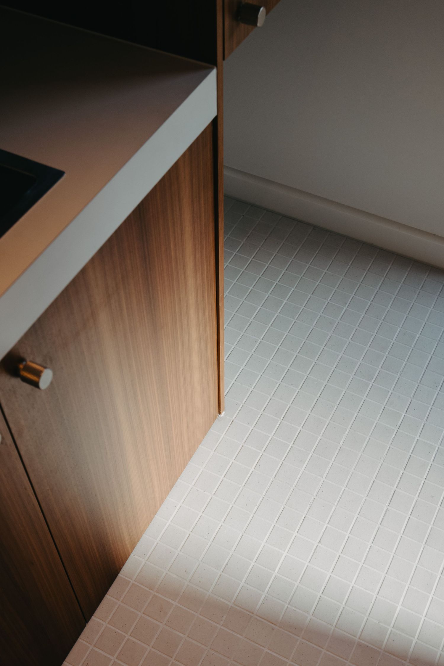 Close-up of walnut kitchen cabinet with brushed brass knob beside white rectangular mosaic tile floor.