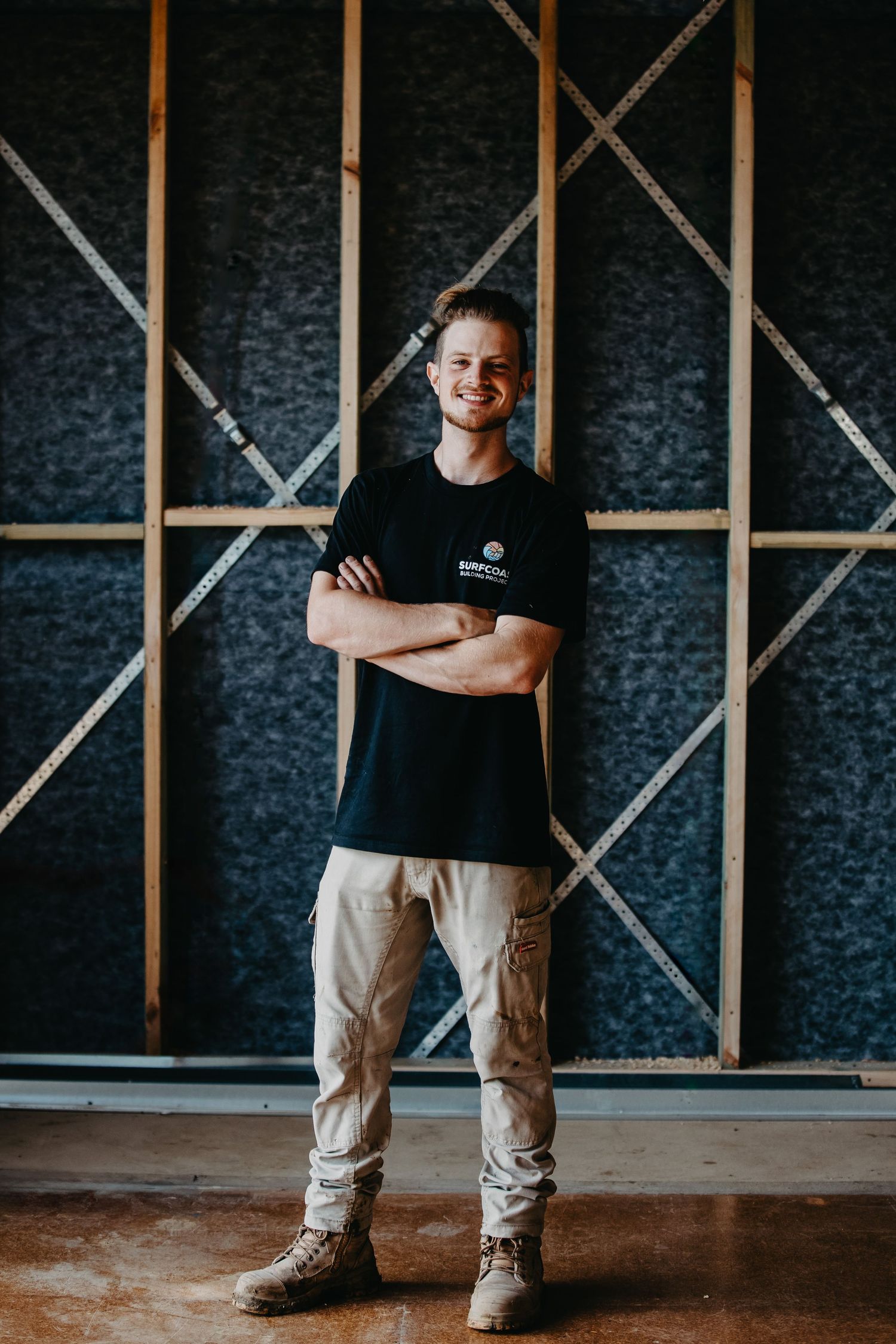 Young man in black t-shirt and beige pants standing with arms crossed smiling in a construction site.