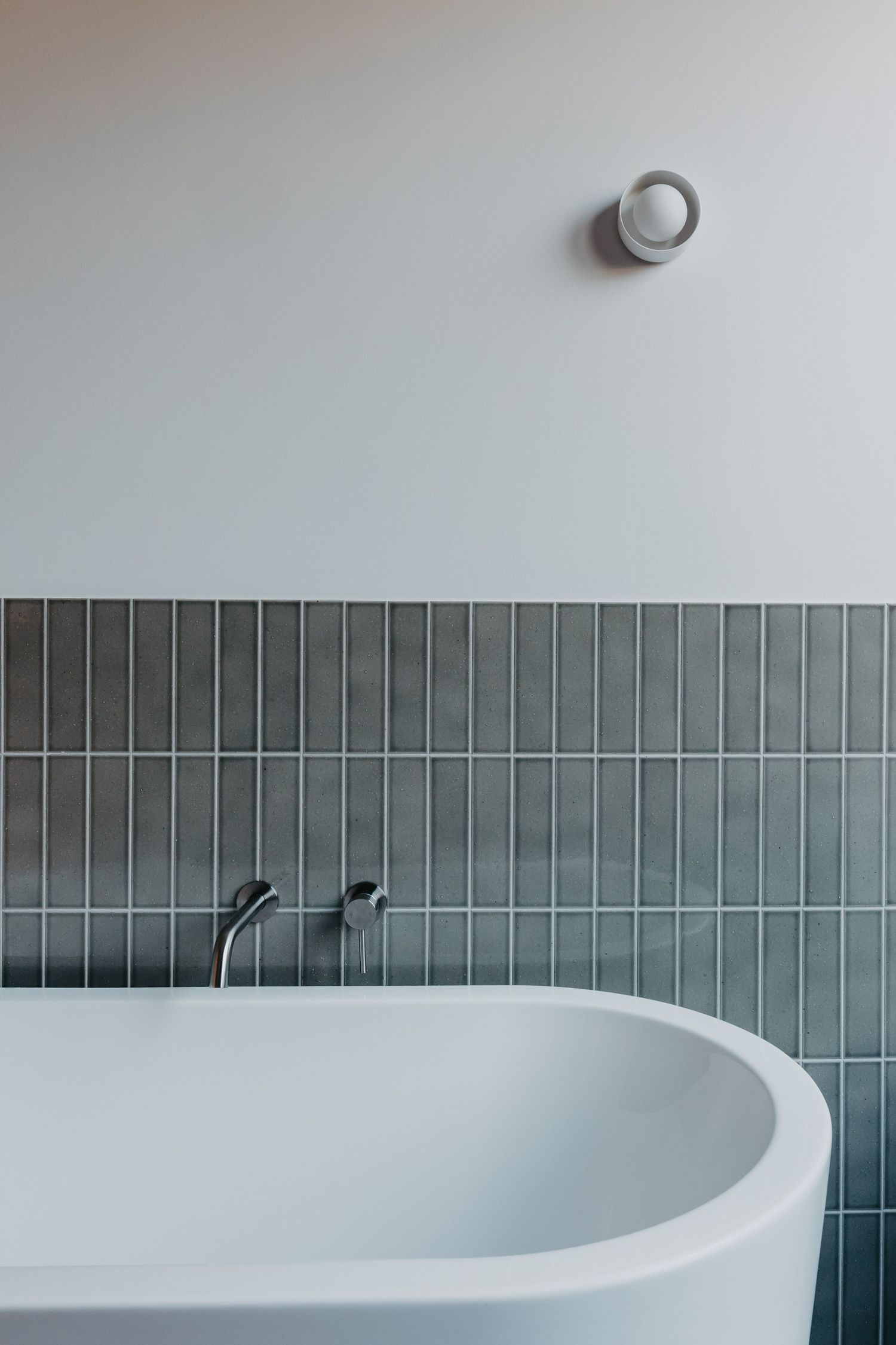 Modern white freestanding bathtub with chrome faucet against green subway tiles and white wall with round sconce light.
