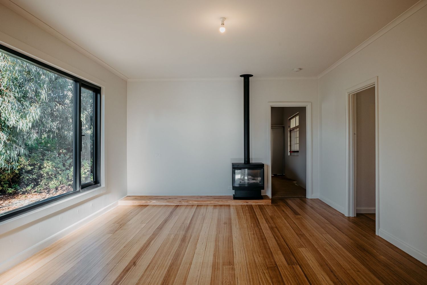 Bright open living room with wide black-framed window, polished timber floors and freestanding black wood fireplace.