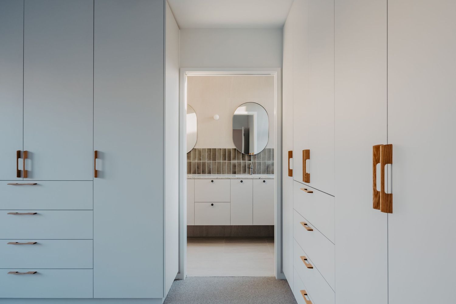 Modern walk-in closet with light blue cabinetry and copper handles leading to a bright bathroom with round mirror.