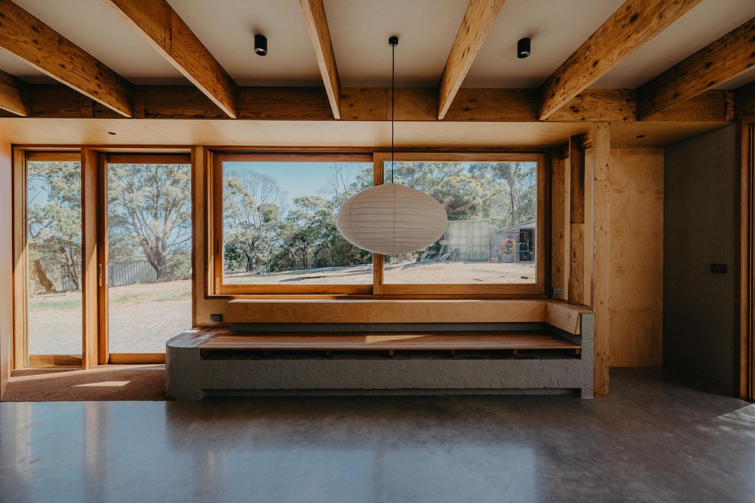 Minimalist interior with polished concrete floor, exposed timber beams, large picture window and hanging Noguchi paper pendant light.