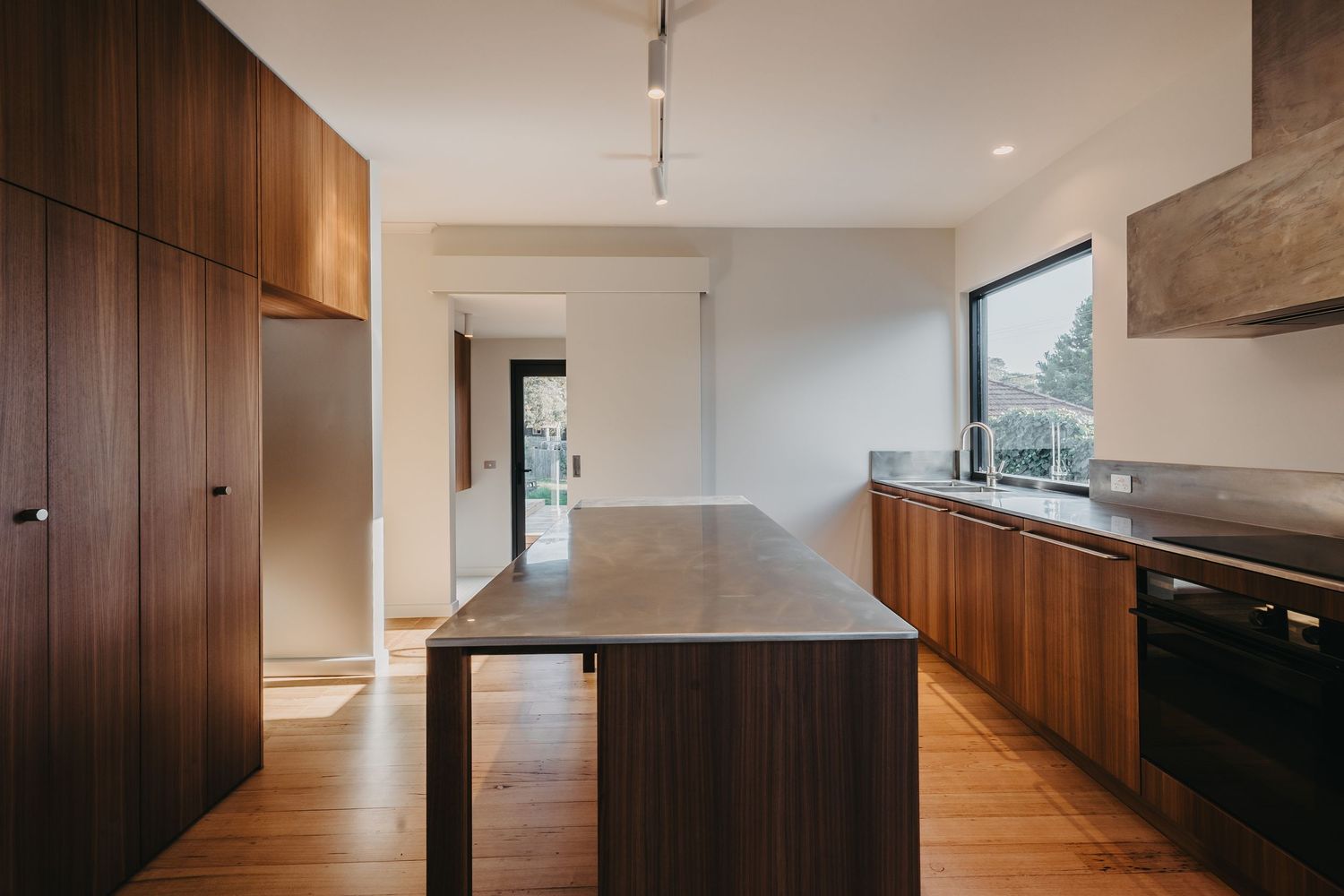 Minimalist kitchen with dark walnut island bench, matching cabinetry, timber floors and black-framed window overlooking garden.