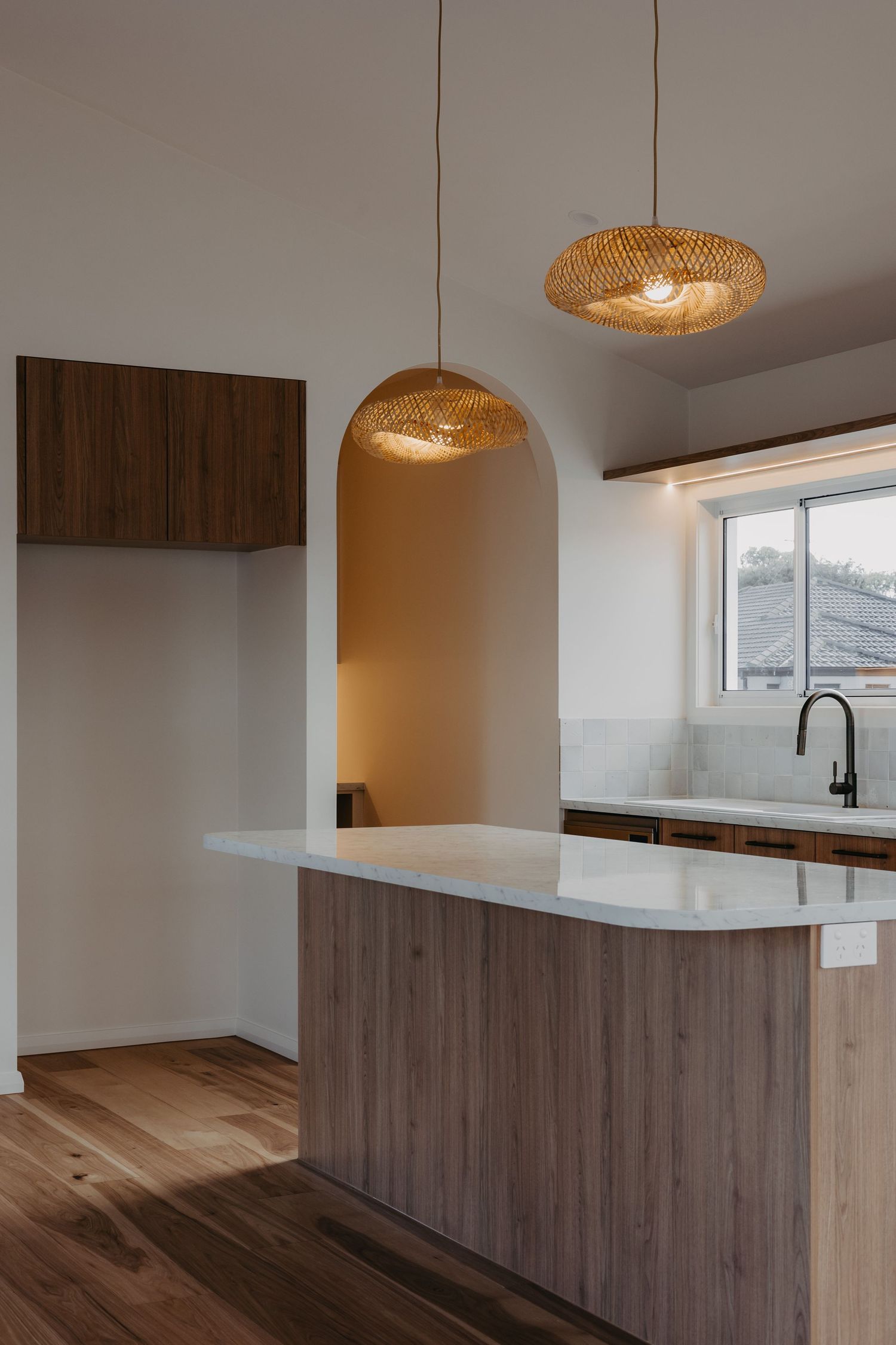 Contemporary kitchen featuring a curved timber island, white benchtop, woven rattan pendant lights, and an arched wall niche.