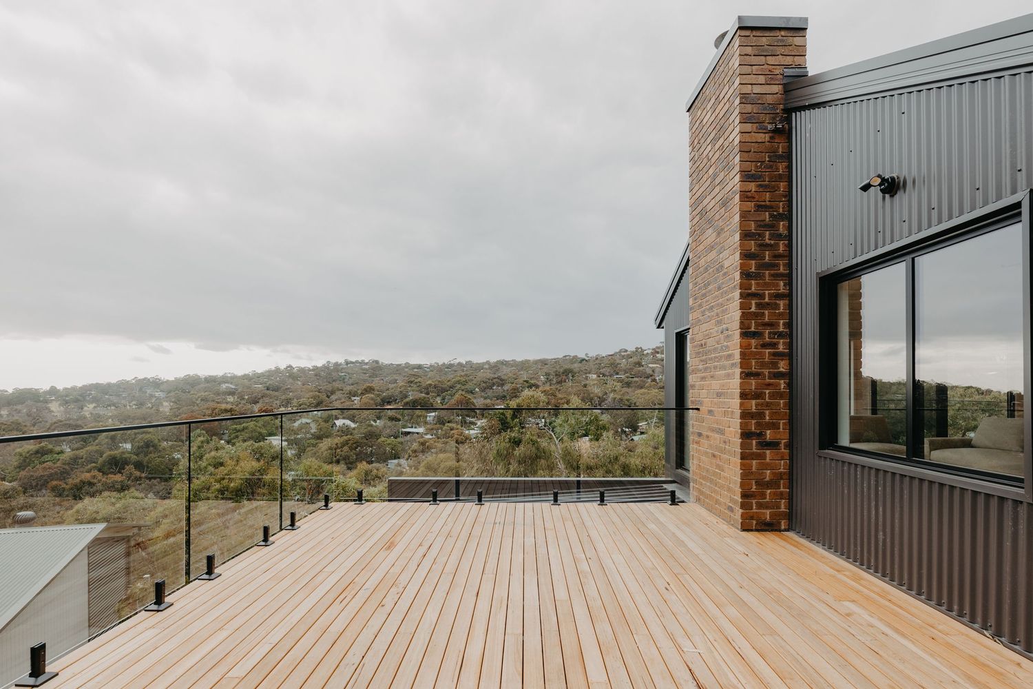 Timber deck with glass balustrade offering panoramic views of rolling hills on a cloudy day.