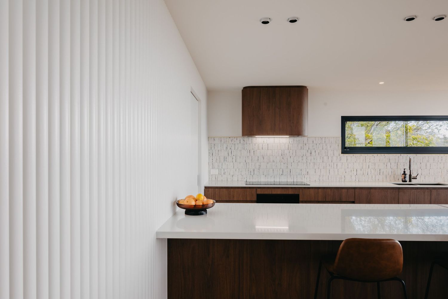 Contemporary kitchen with walnut cabinetry, white quartz countertop island, textured white backsplash tiles and integrated range hood.