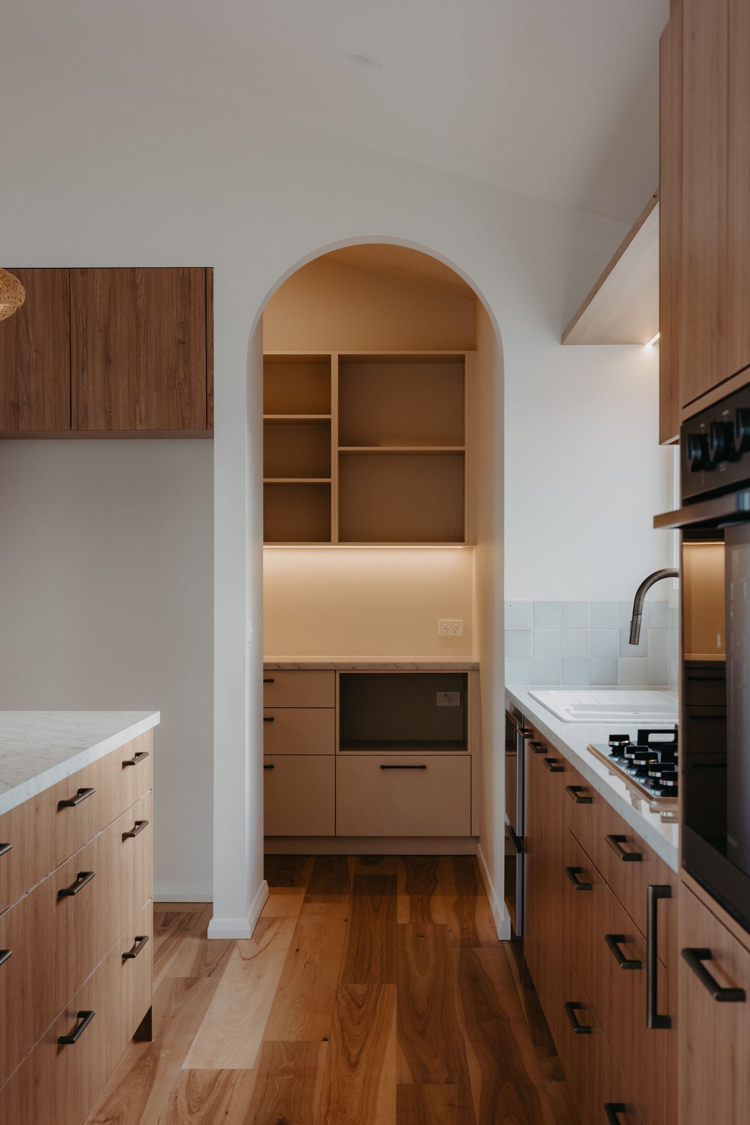 Kitchen with warm wood cabinetry, arched pantry alcove featuring open shelving and microwave, black handles, and light hardwood floors.