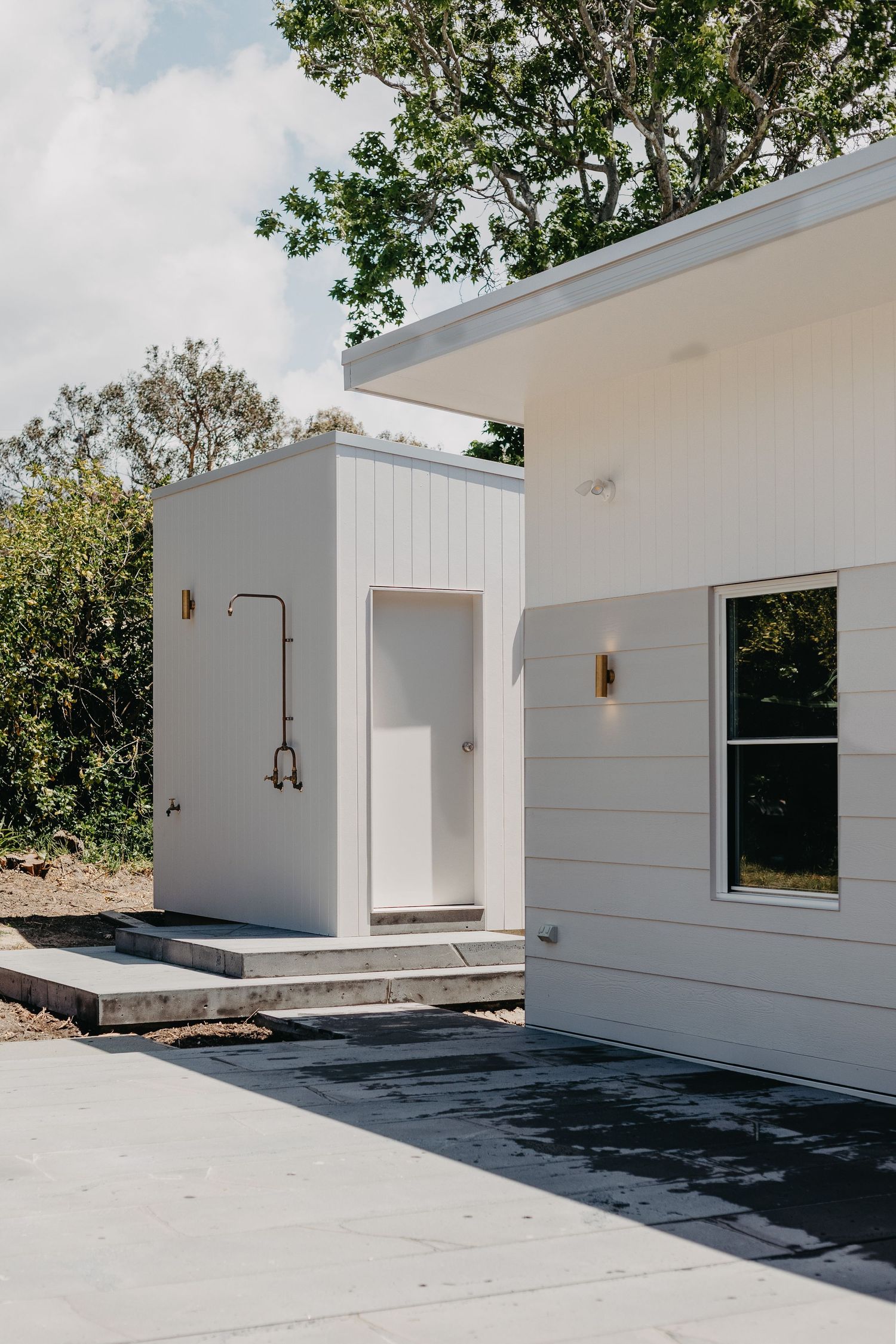Modern white farmhouse exterior with outdoor shower, board and batten siding, brass fixtures, and lush green trees.