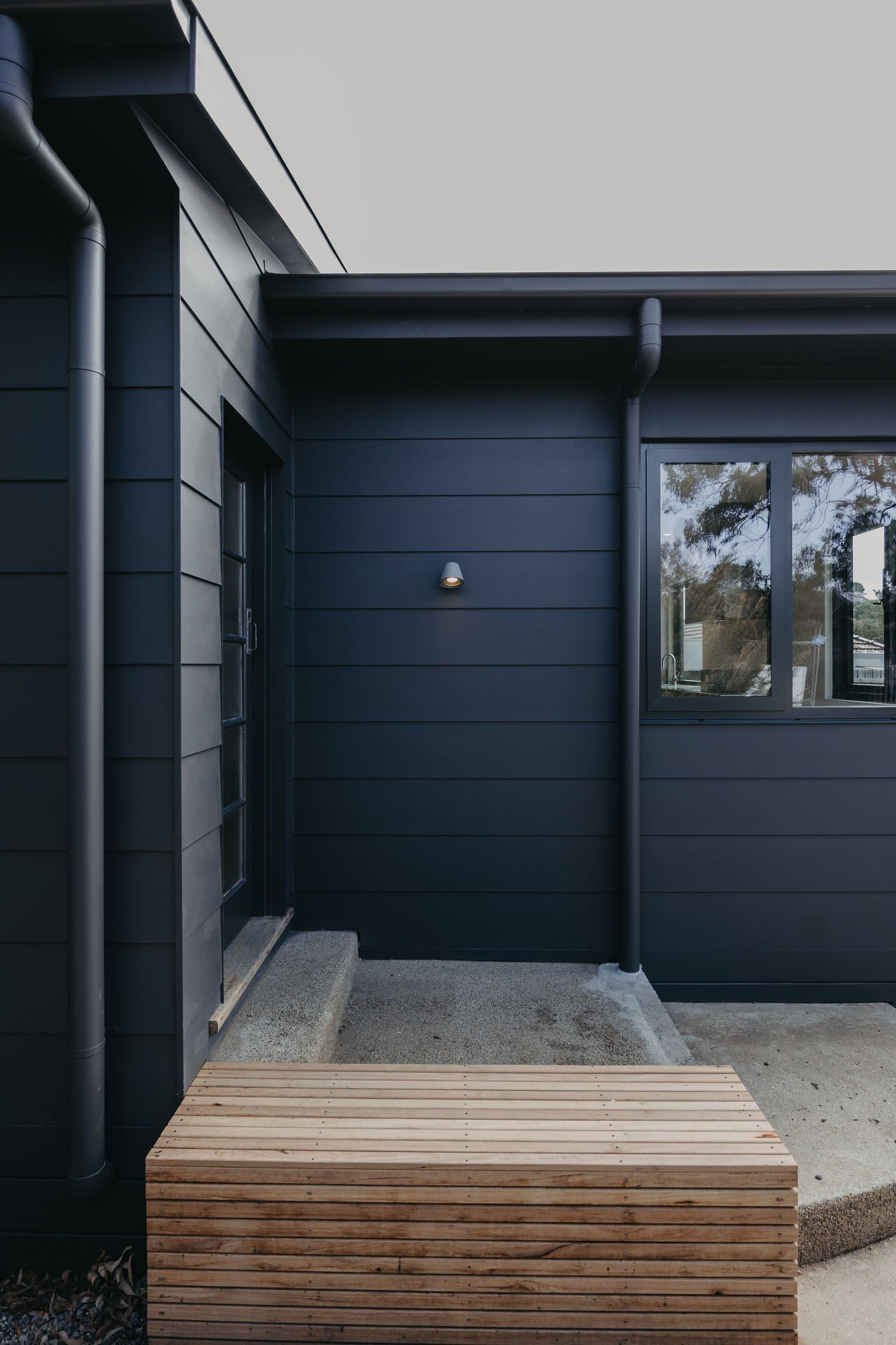 Dark navy blue exterior cladding of a modern home with timber deck step and black framed windows.