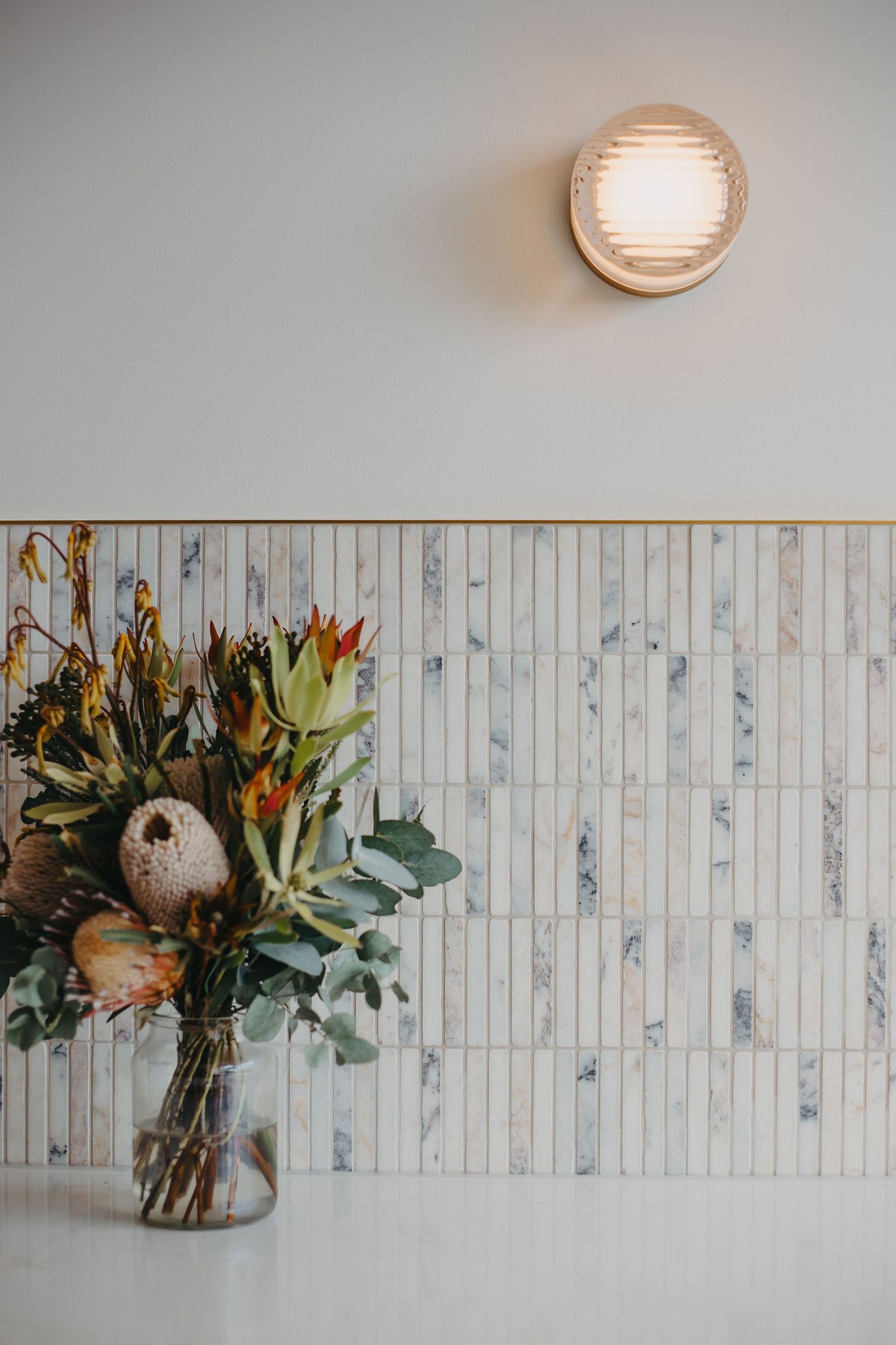 Decorative native flower bouquet in glass vase against marble tile backsplash with gold trim and wall sconce.