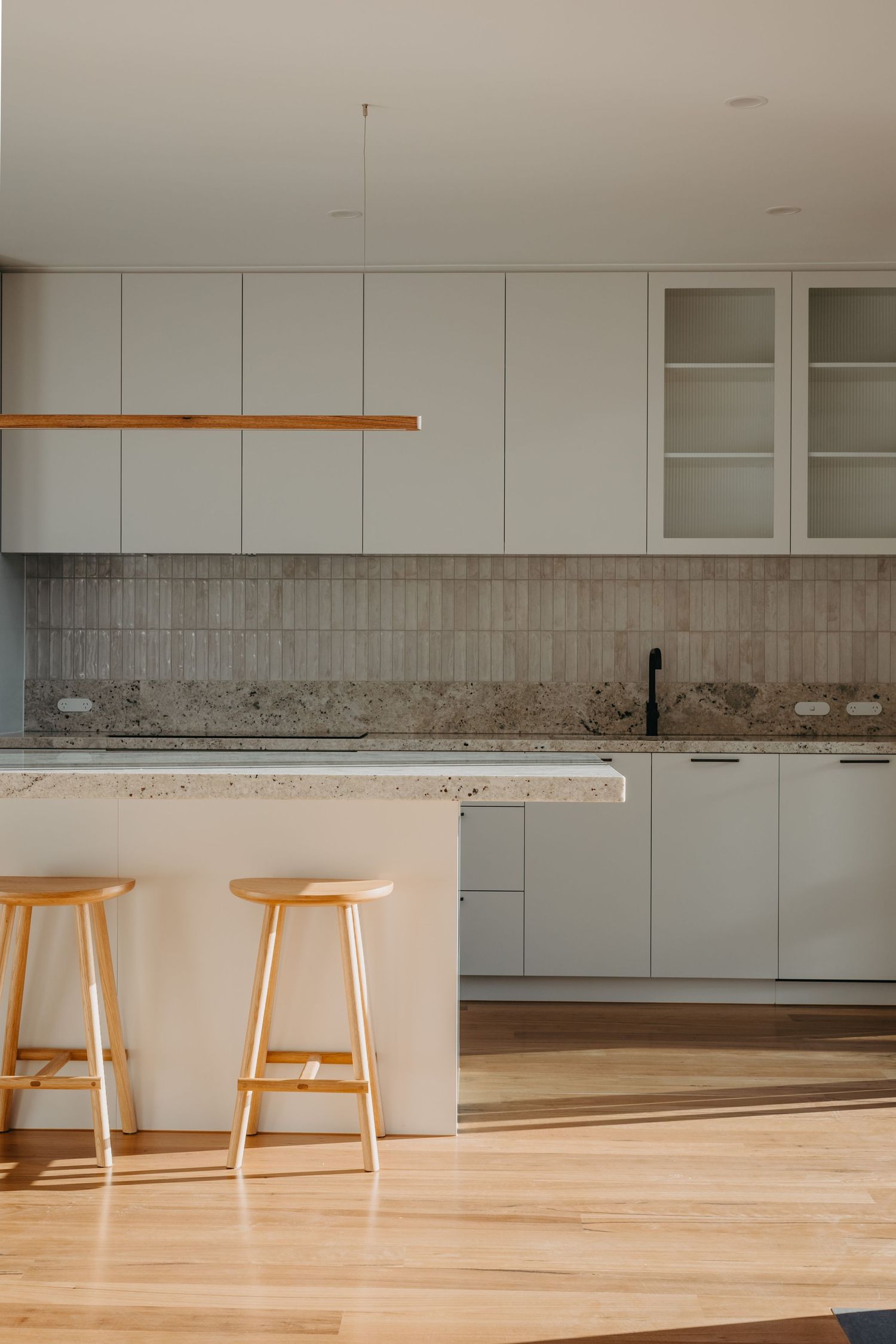 Modern white kitchen with granite island, two wooden bar stools, tile backsplash, and warm hardwood floors.