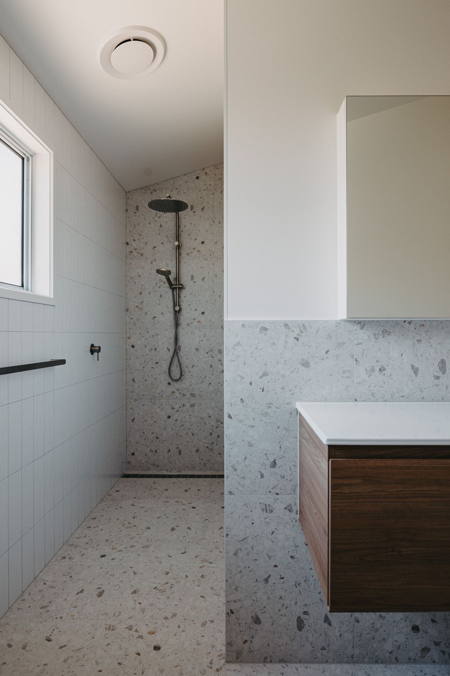 Modern bathroom with terrazzo tiles, walk-in shower, dark fixtures, floating timber vanity, and white walls.