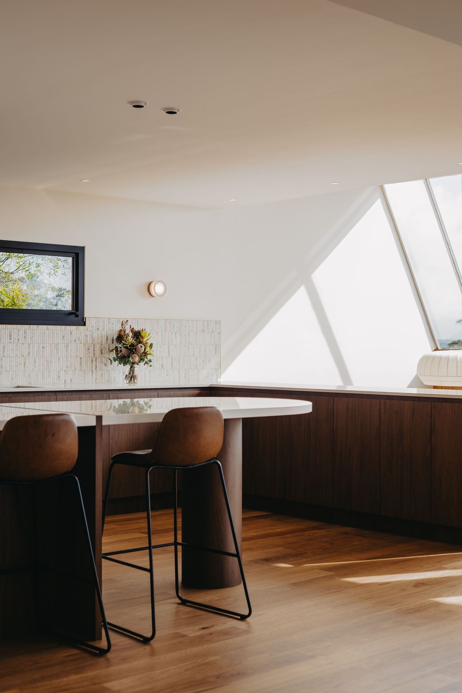 Sun-drenched modern kitchen with walnut island, leather stools, fresh flowers, and dramatic skylight window.