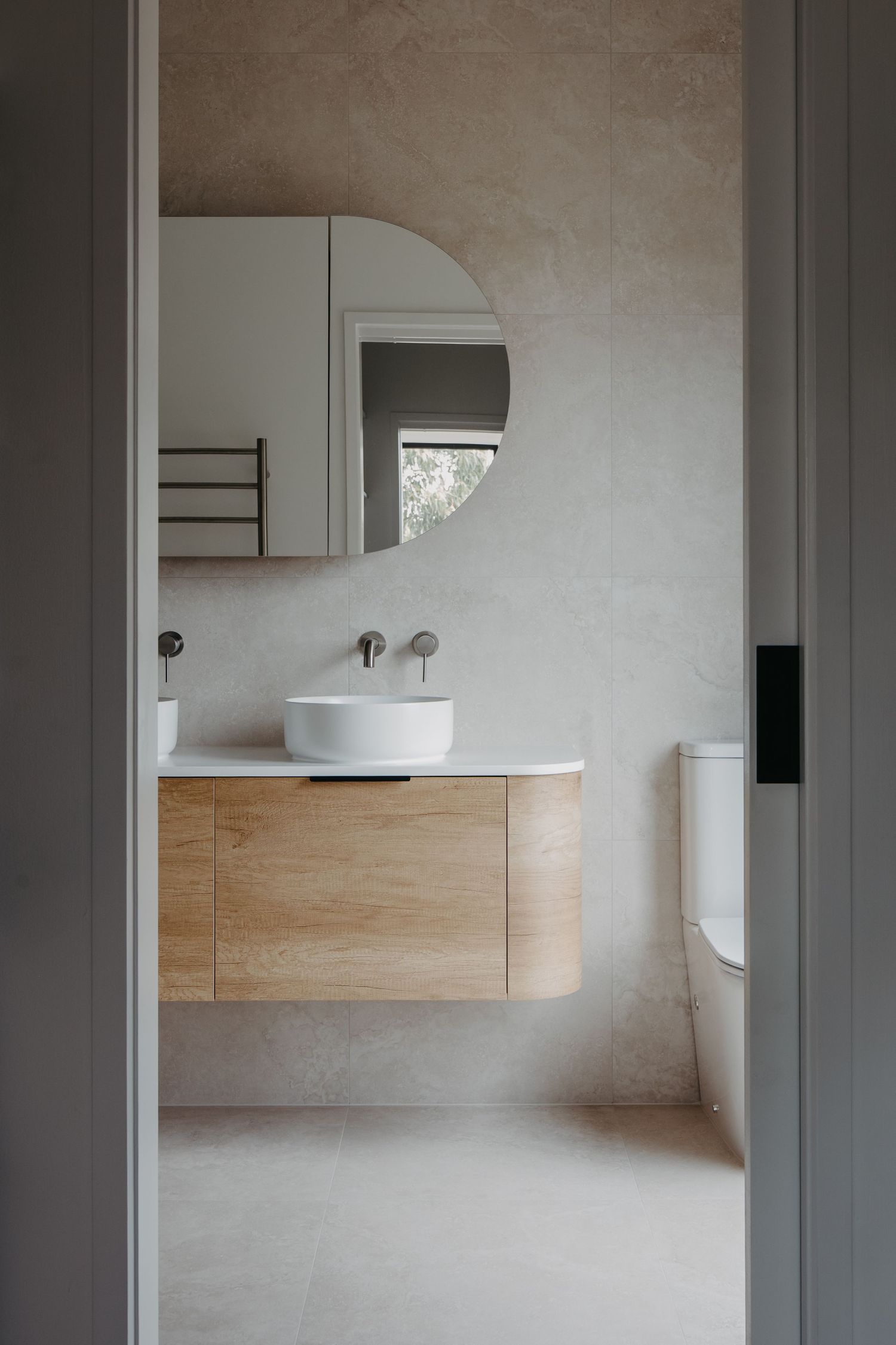 Minimalist bathroom with timber vanity, round vessel sink, arched mirror, and textured concrete-look walls.